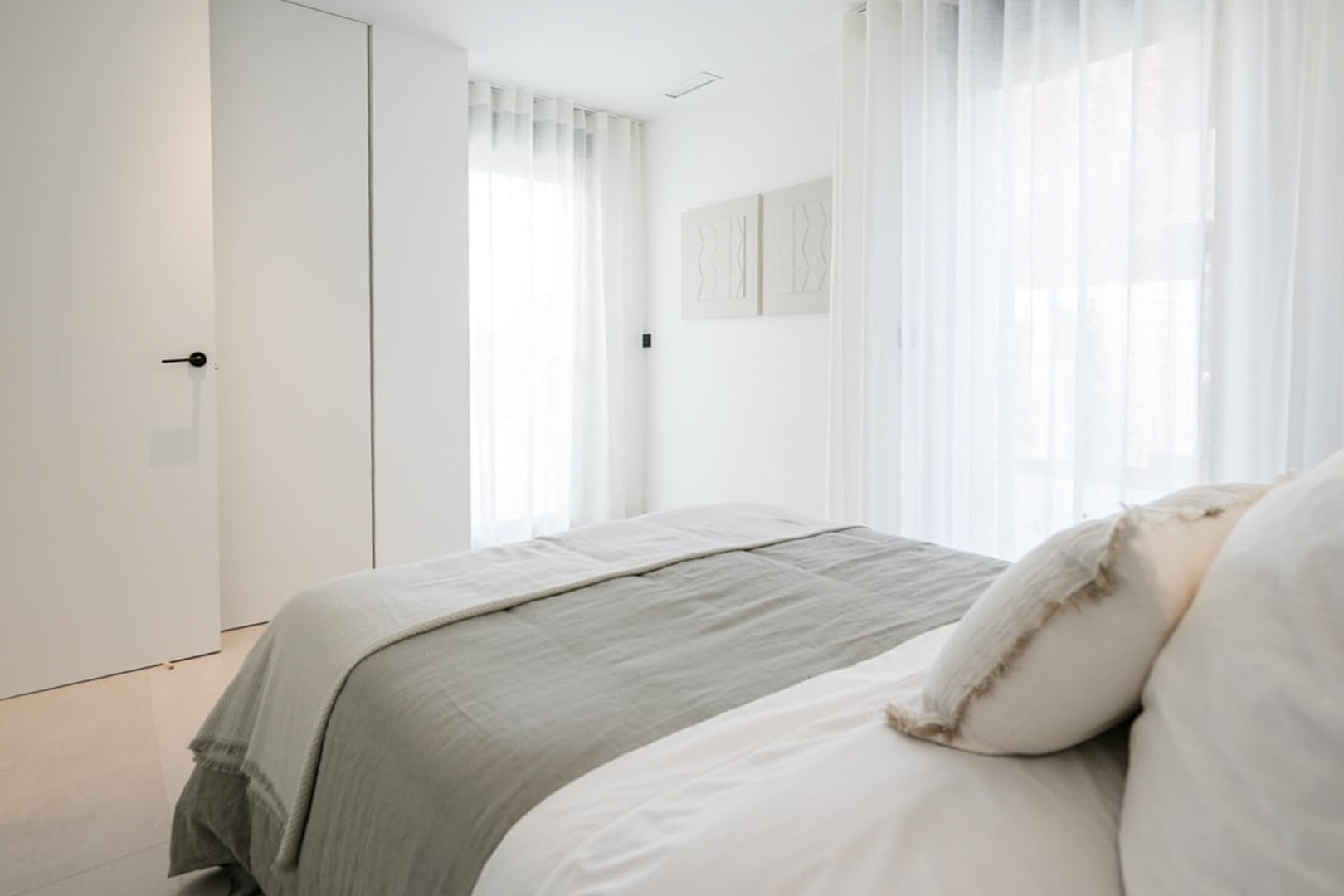 Chambre lumineuse avec literie grise et blanche, rideaux transparents et décoration murale minimaliste.