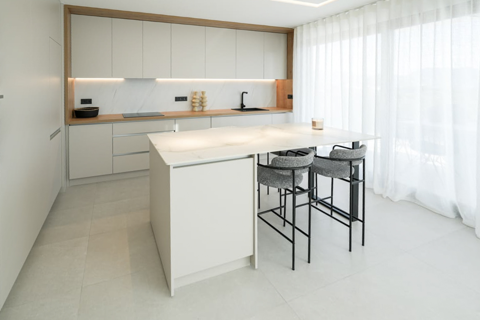 Cuisine moderne avec îlot en marbre, tabourets gris et armoires blanches sous lumière naturelle.