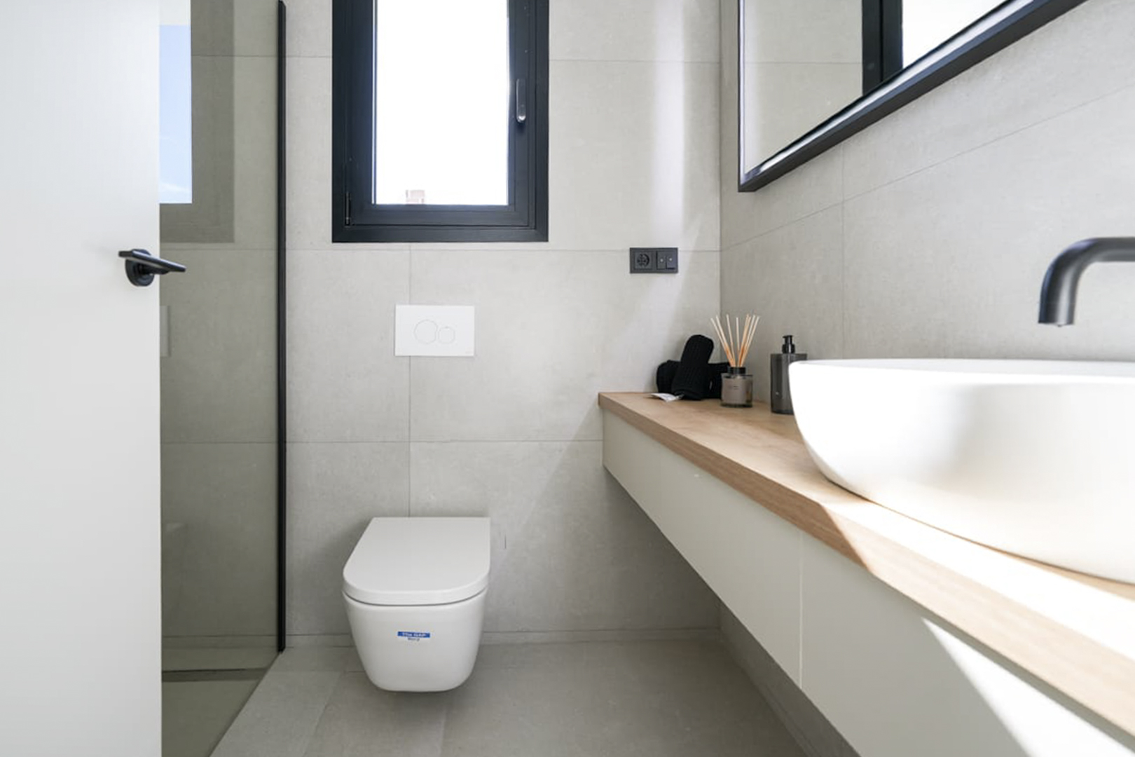 Détail d'une salle de bain moderne avec comptoir en bois clair, lavabo blanc et carrelage gris neutre.