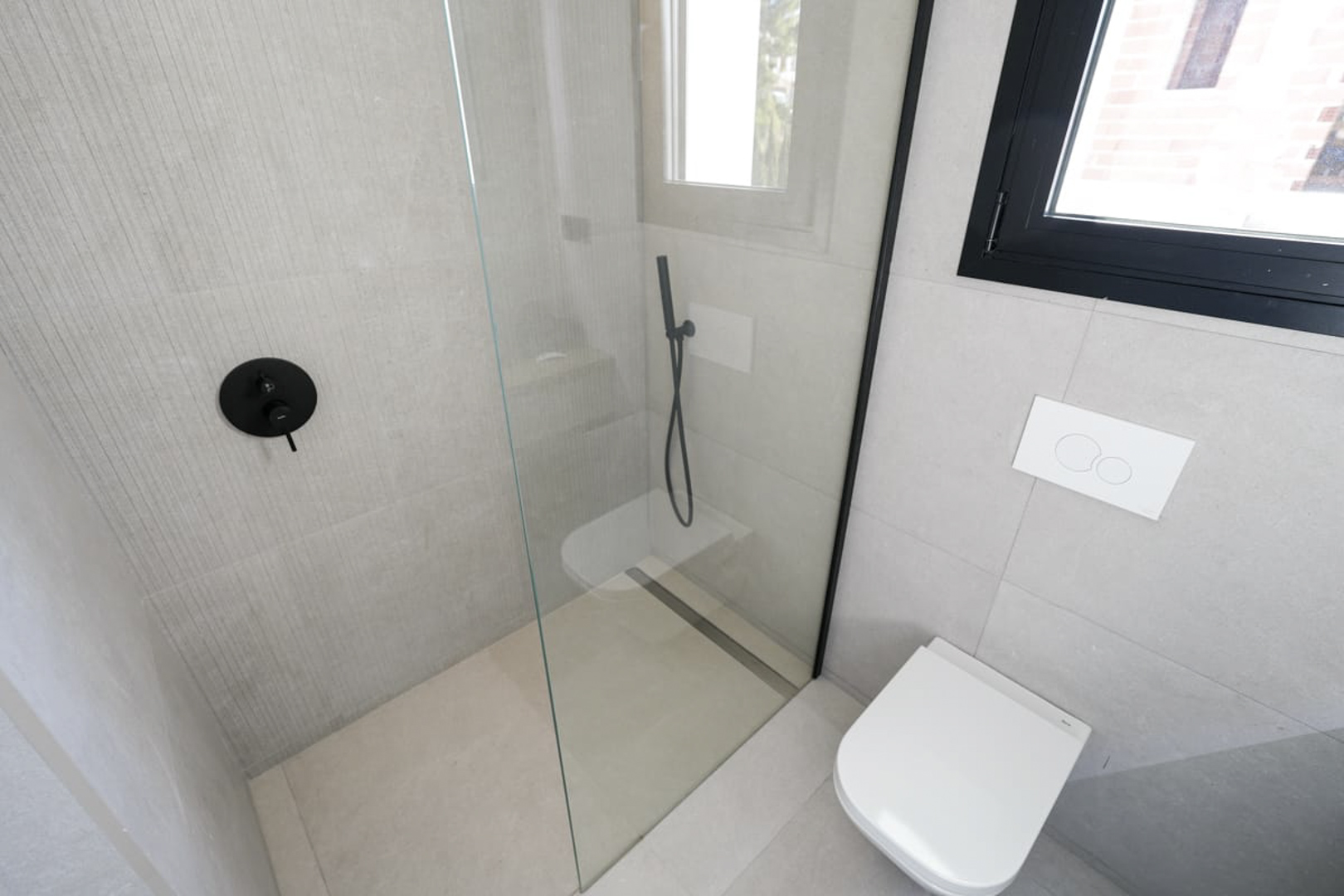 Détail d'une salle de bain moderne avec douche en verre, robinetterie noire et carrelage gris texturé.