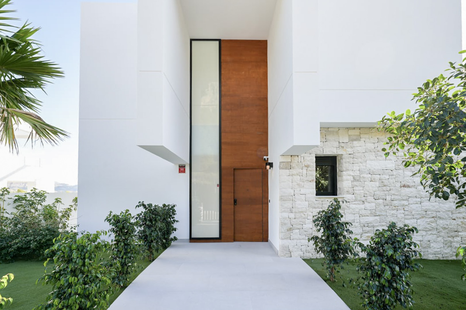 Façade moderne avec porte en bois et détails en pierre claire, entourée de verdure.