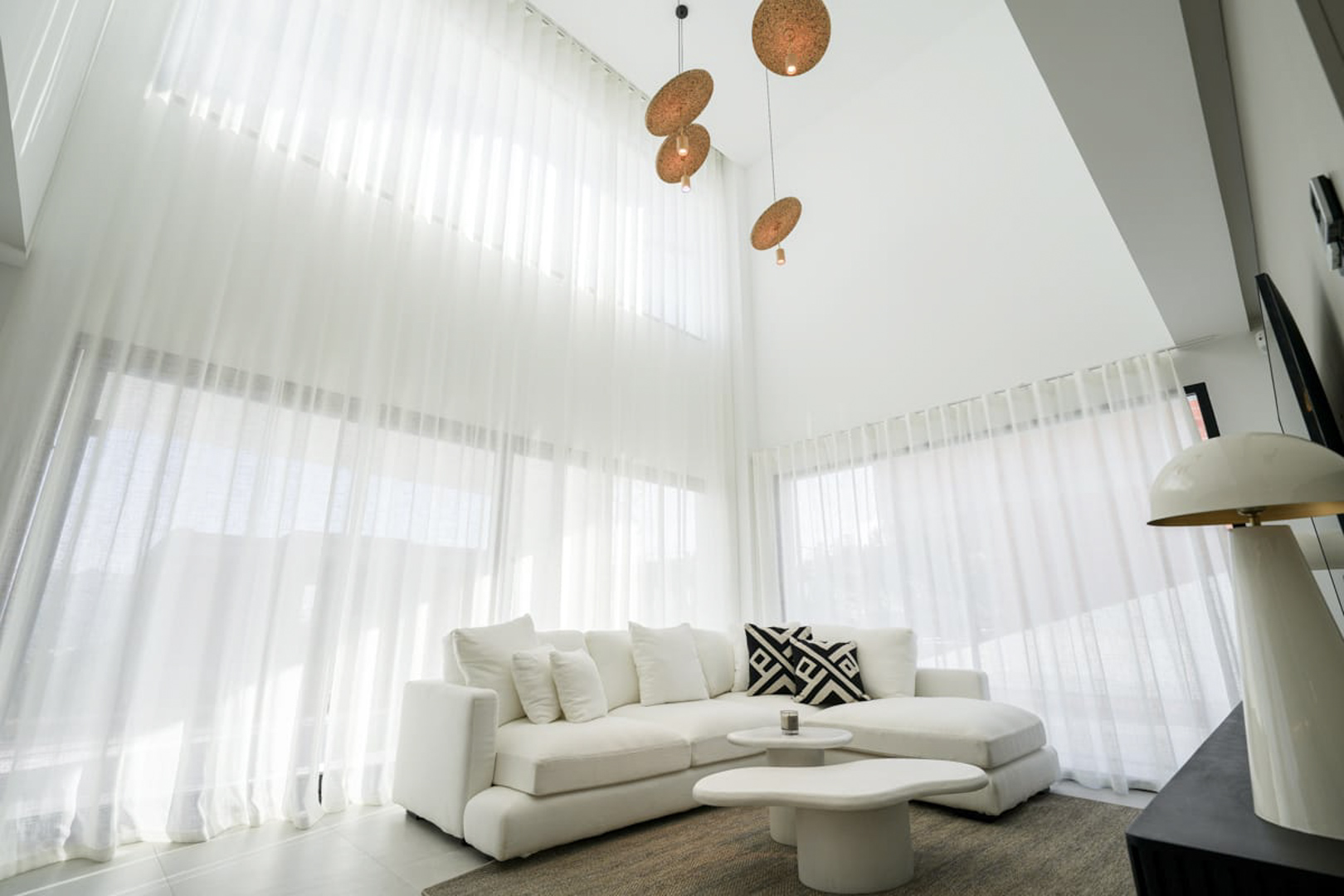 Salon lumineux avec rideaux blancs, canapé d'angle et suspensions design, créant une ambiance moderne et minimaliste.