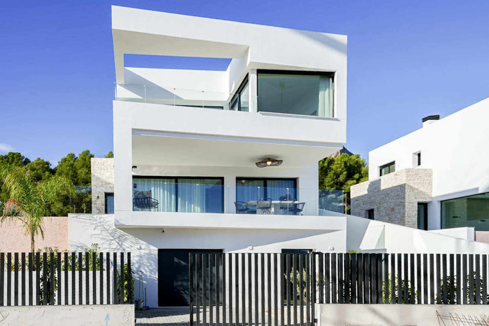 Vue extérieure d'une villa moderne de trois étages avec balcons, garde-corps en verre et clôture à lattes noires.