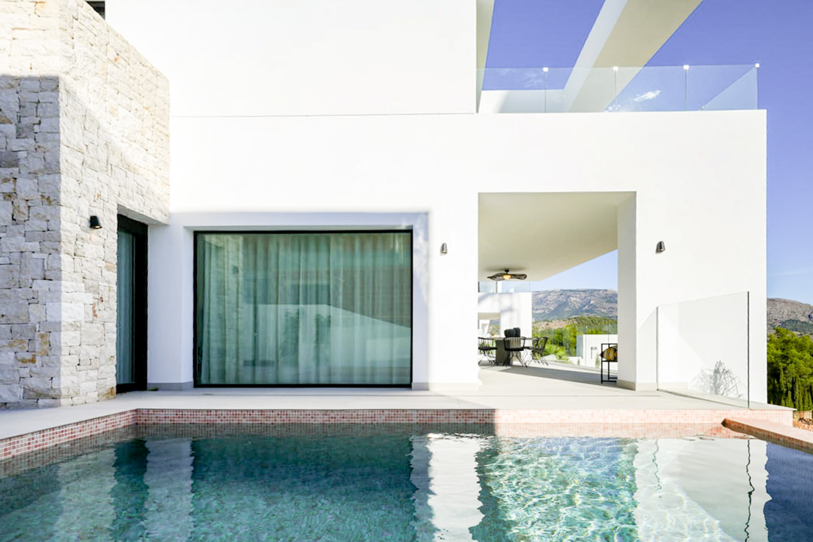 Vue sur la piscine avec bordure en mosaïque, façade en pierre et terrasse couverte avec salle à manger extérieure.