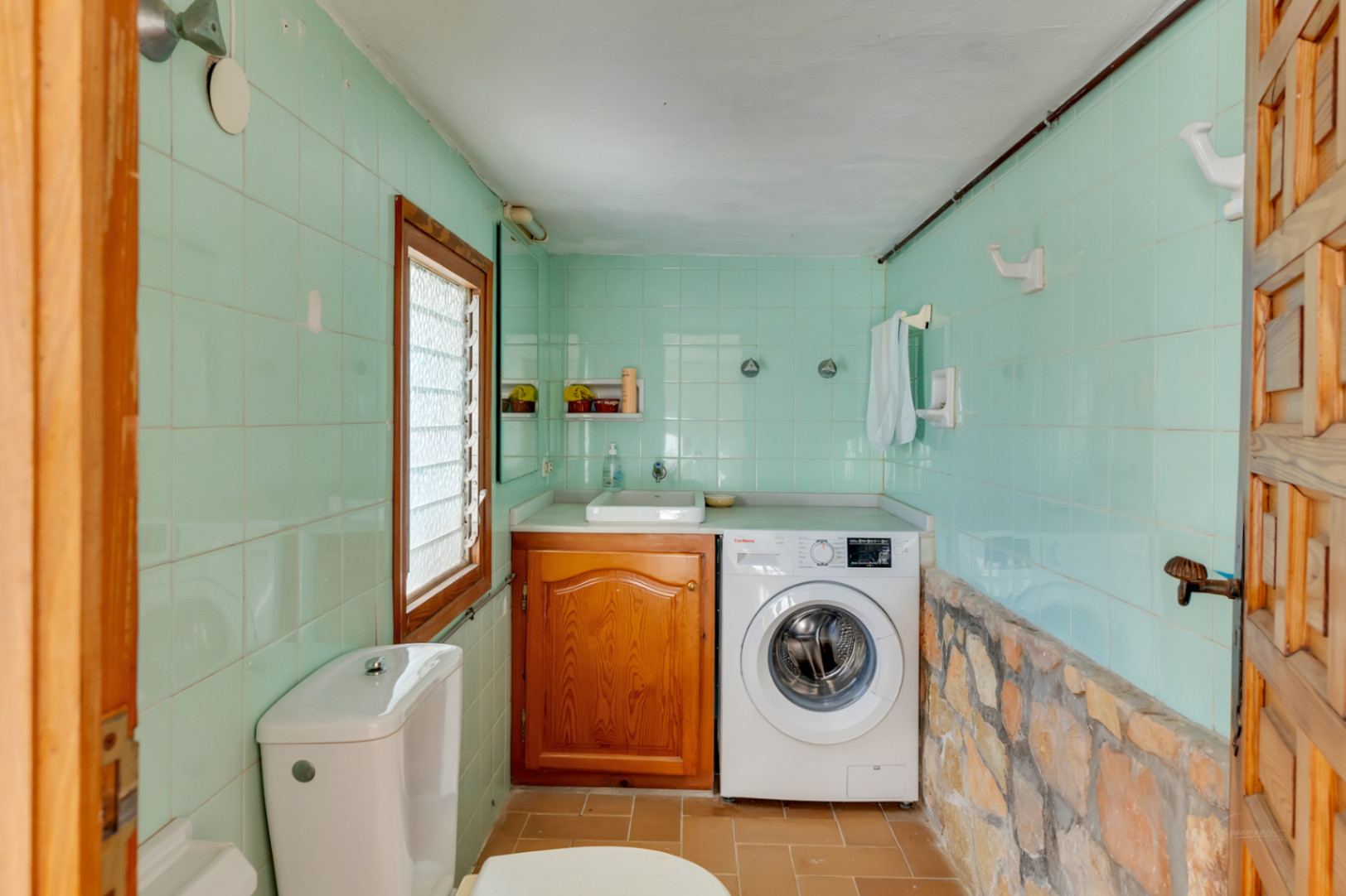Buanderie et toilettes avec carrelage vert menthe, mur en pierre naturelle, meuble en bois sous vasque et lave-linge intégré.