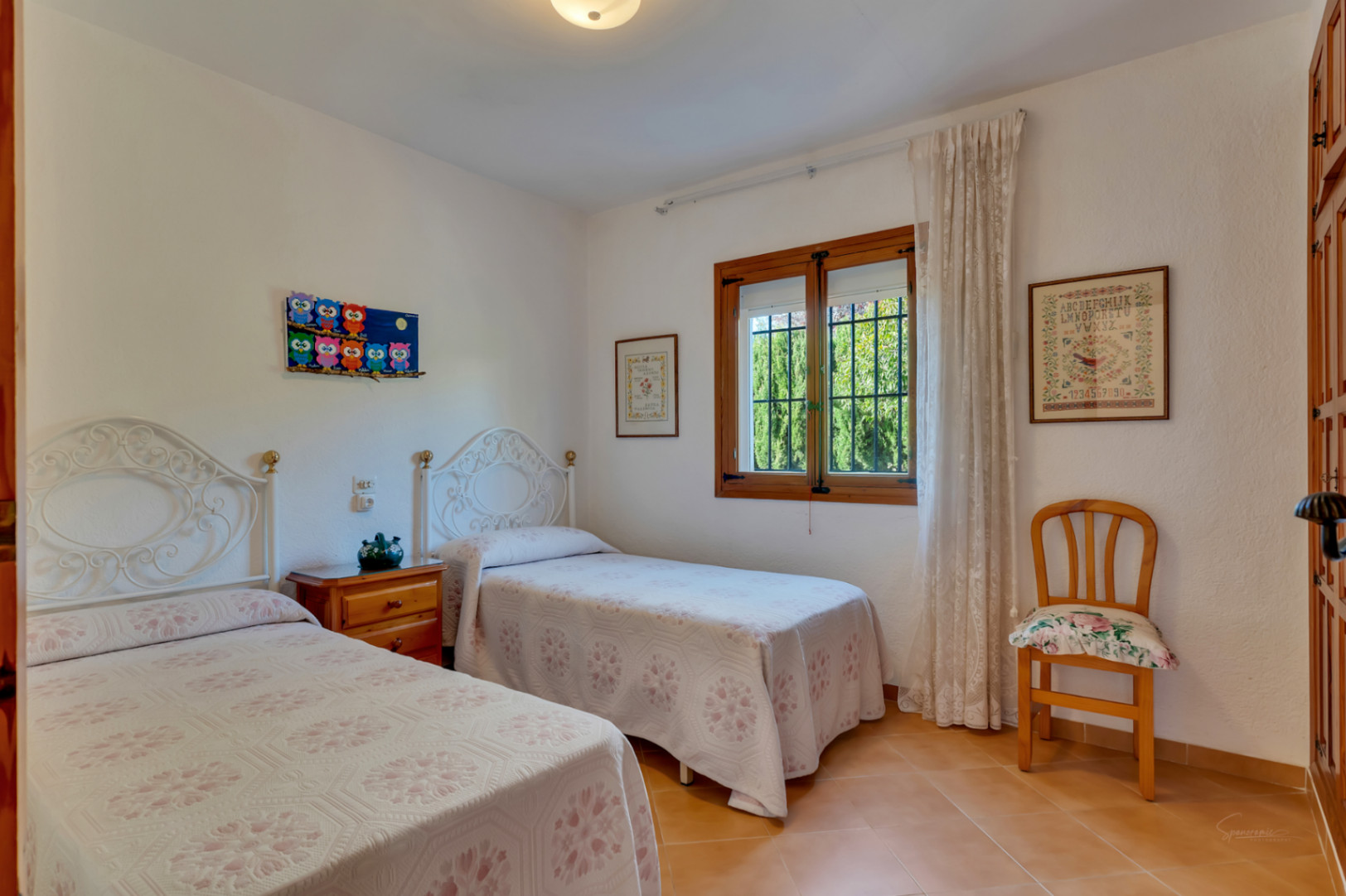 Chambre lumineuse avec deux lits simples, têtes de lit en fer forgé blanc, sol en terre cuite et fenêtre en bois traditionnelle.
