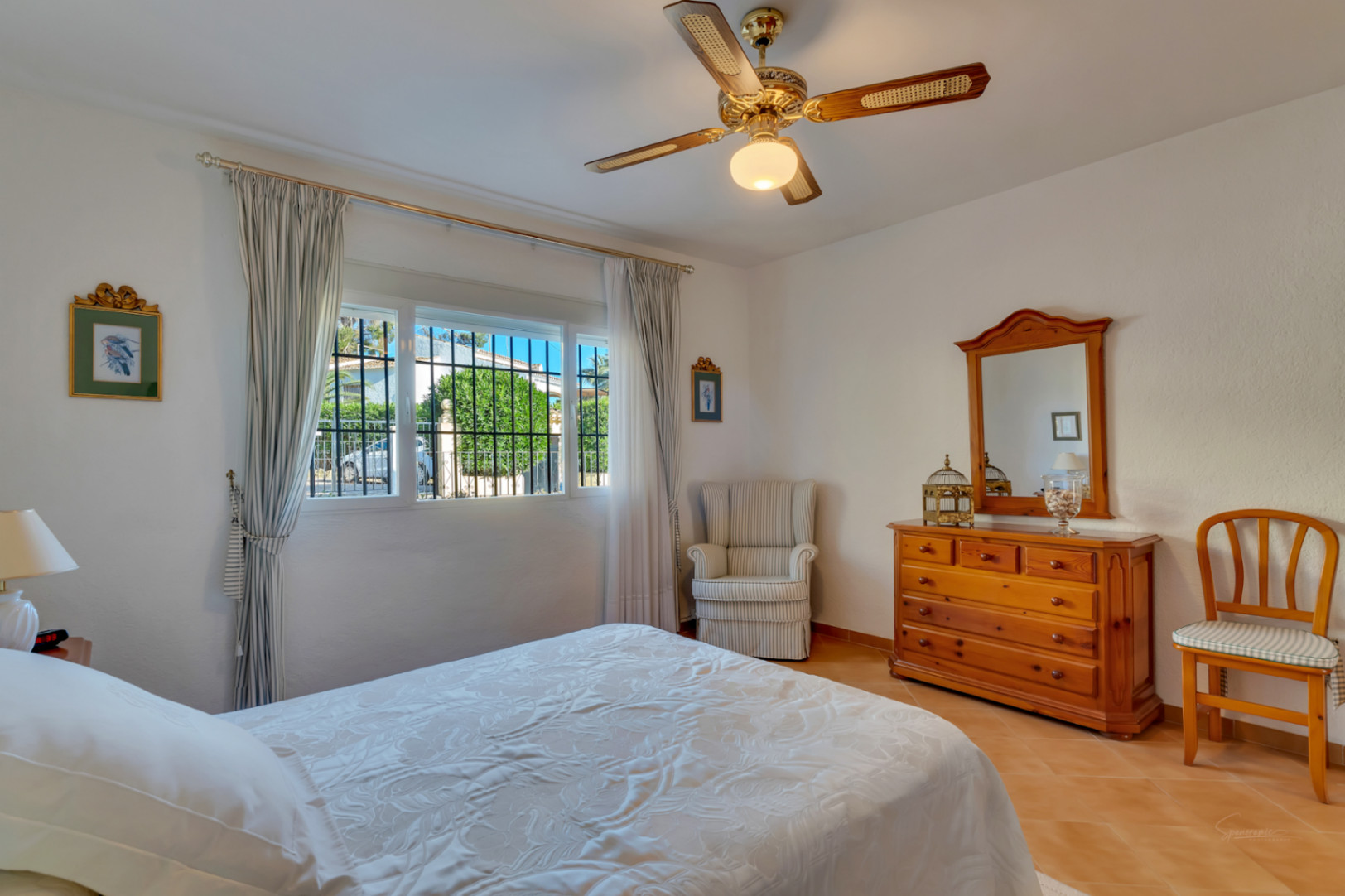 Chambre lumineuse avec sol en terre cuite, mobilier en bois traditionnel, ventilateur de plafond et fenêtre offrant une vue sur le jardin.