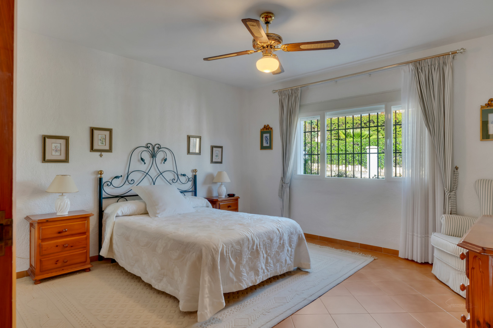 Chambre lumineuse avec tête de lit en fer forgé, sol en terre cuite et ventilateur de plafond. La fenêtre sécurisée offre une vue dégagée sur la végétation du jardin.