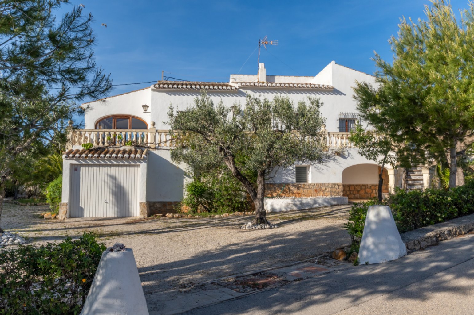 Façade de villa méditerranéenne avec murs blancs, terrasse à balustrade et garage. L'extérieur dispose d'un olivier central et d'un soubassement en pierre naturelle.