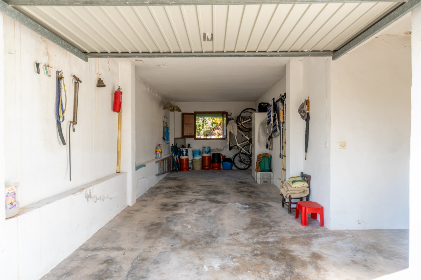 Grand garage fermé avec sol en béton et murs blancs, offrant un vaste espace de rangement et une ventilation naturelle par une fenêtre au fond.