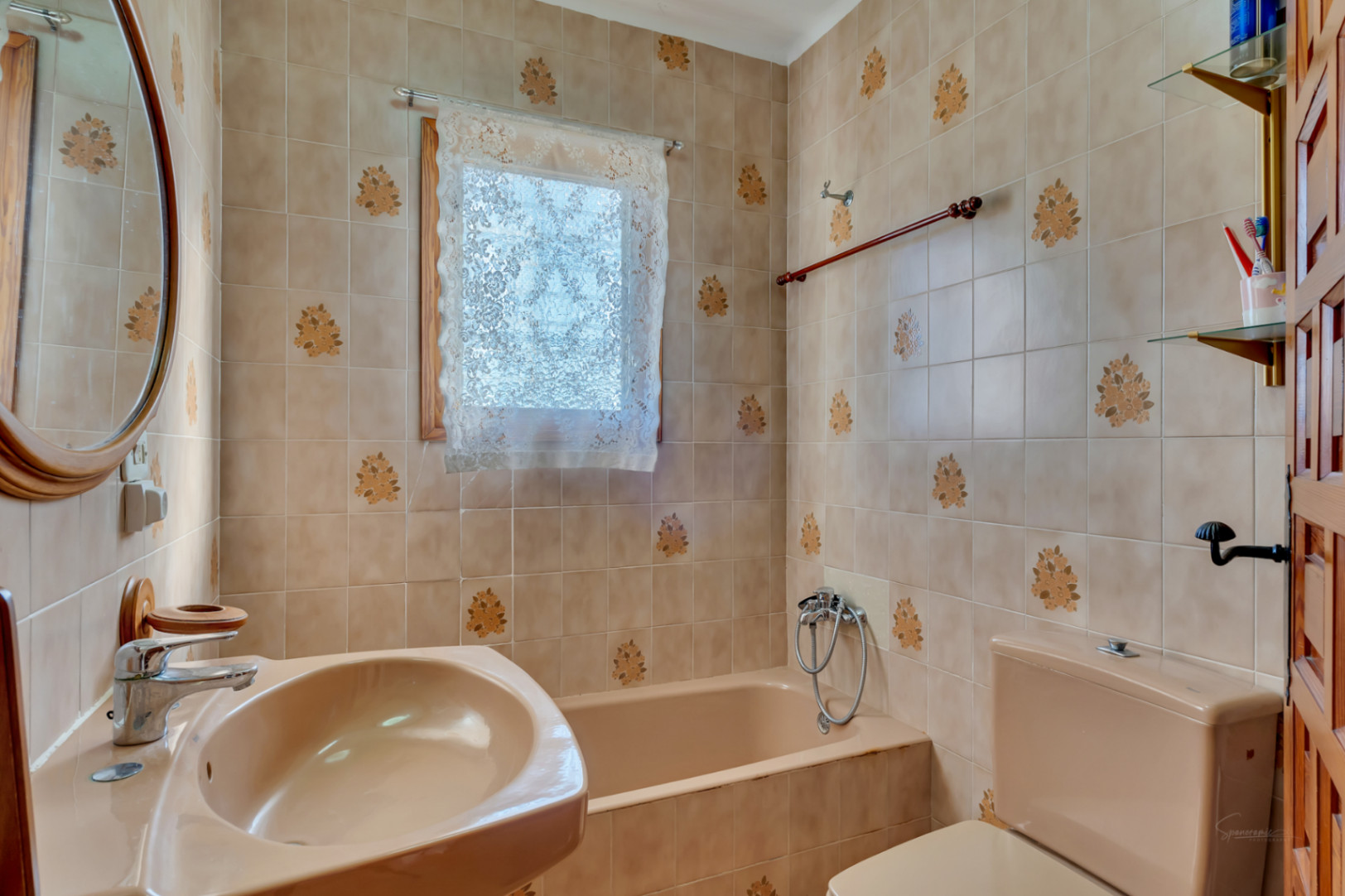 Salle de bain de style traditionnel avec carrelage beige à motifs floraux, baignoire intégrée et lavabo assorti, complétée par un miroir ovale et des finitions en bois.