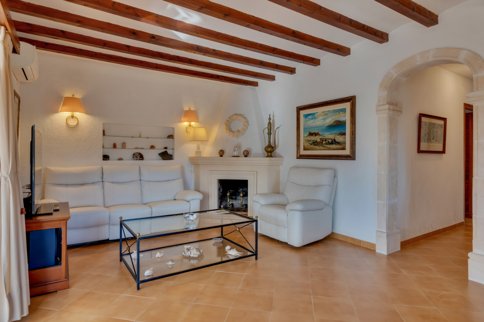 Salon avec poutres en bois apparentes, cheminée classique et arche traditionnelle en pierre Tosca. Équipé de canapés en cuir blanc et d'un sol en grès cérame.