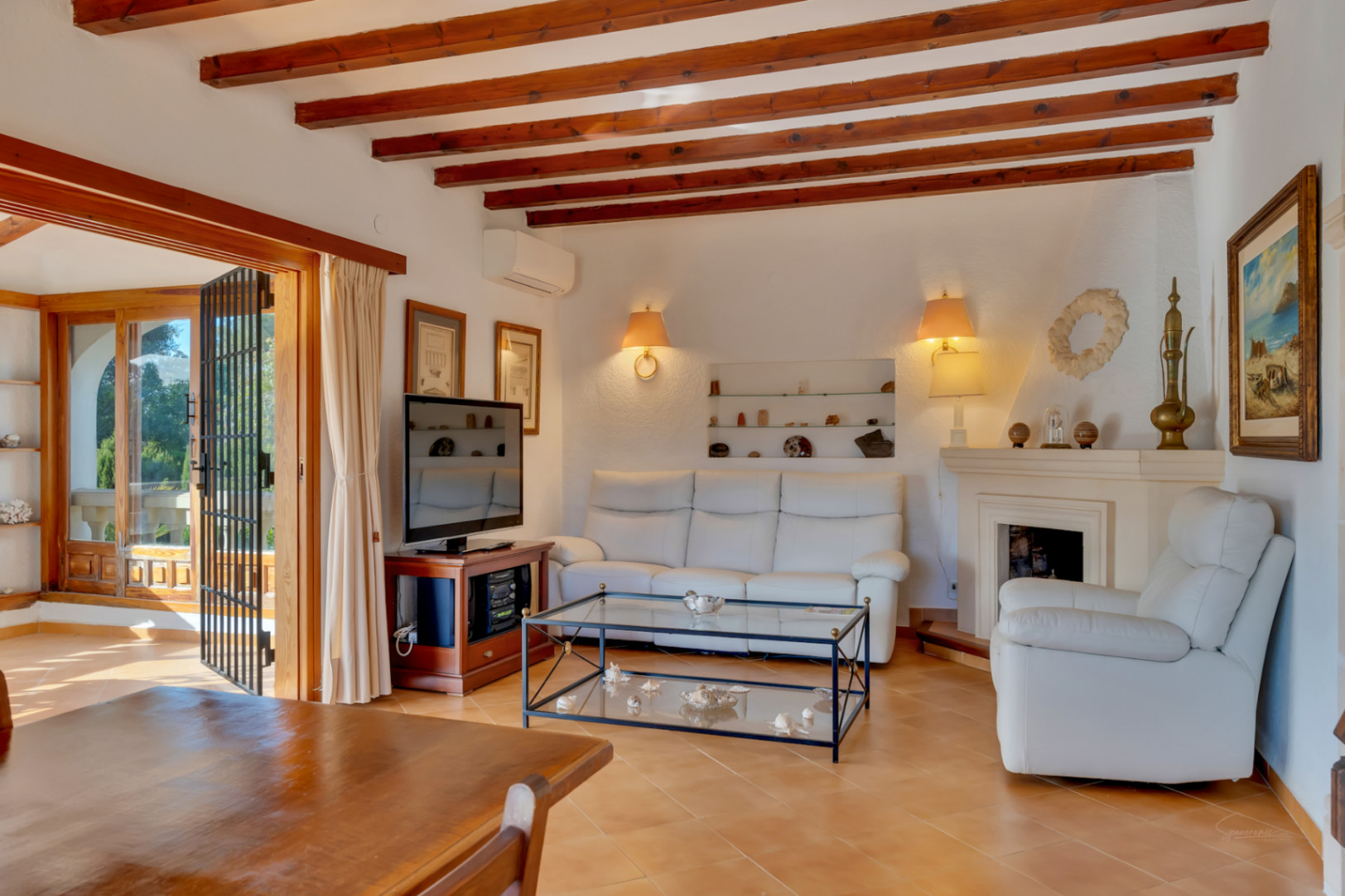 Salon avec poutres en bois apparentes, cheminée classique et canapés en cuir blanc. L'espace offre un accès direct à la terrasse et un sol en grès cérame.