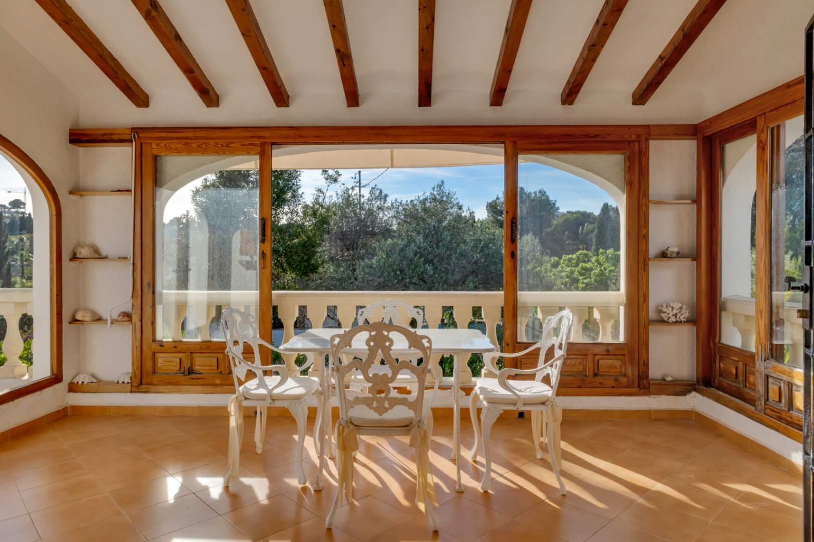 Terrasse couverte vitrée avec poutres en bois apparentes, mobilier de jardin blanc et vue sur la végétation environnante par de larges baies vitrées.