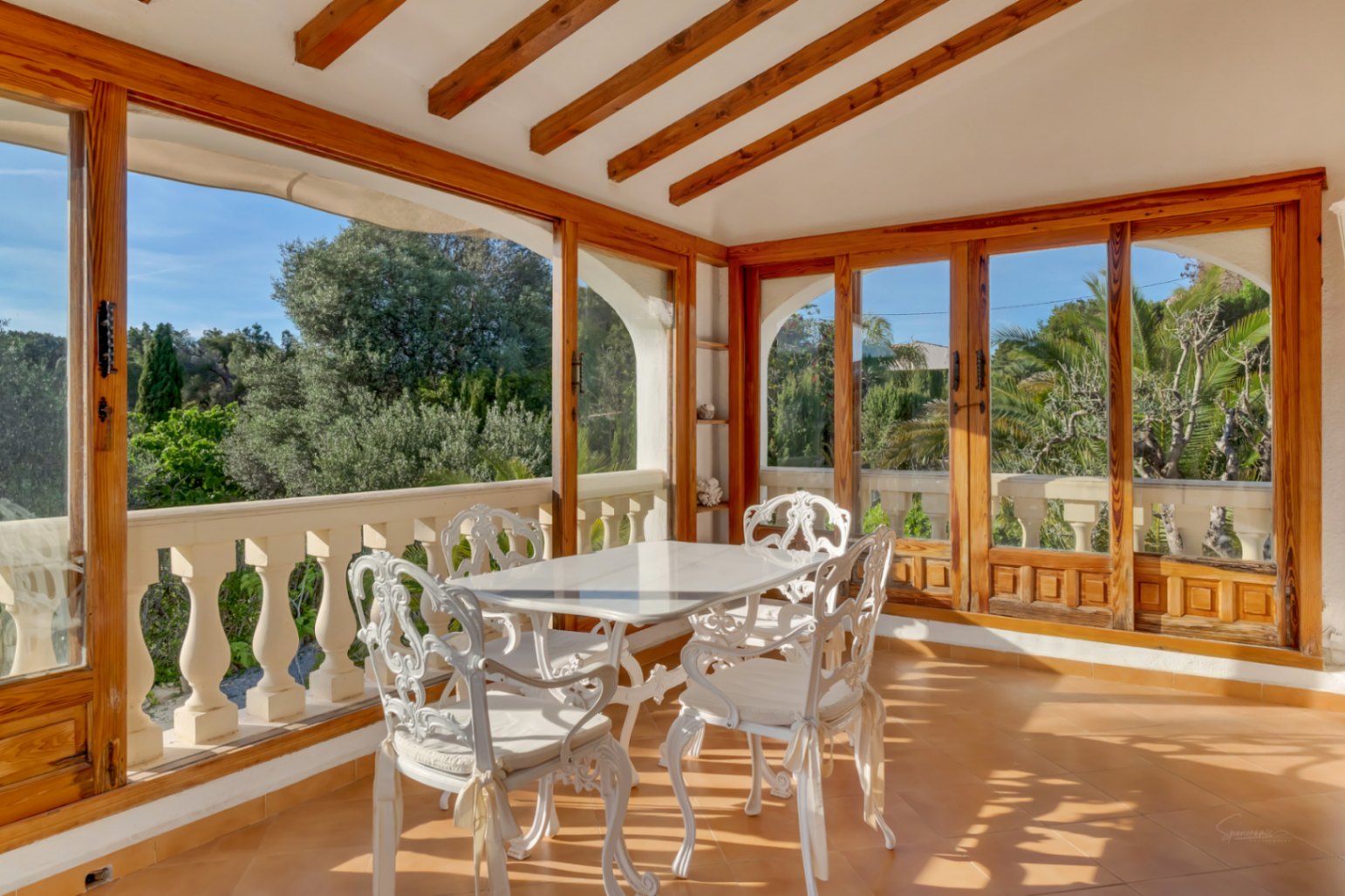 Terrasse vitrée avec poutres en bois apparentes, mobilier en fer forgé blanc et grandes baies vitrées offrant une vue sur le jardin.