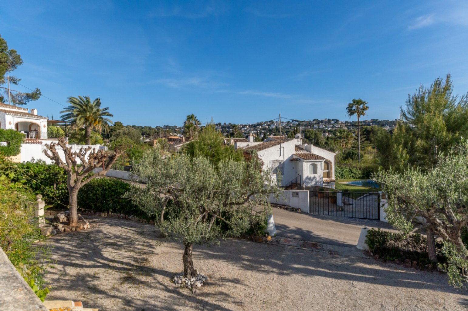 Vue extérieure surélevée montrant le jardin de gravier avec des oliviers et l'architecture traditionnelle de la villa sous un ciel bleu dégagé.