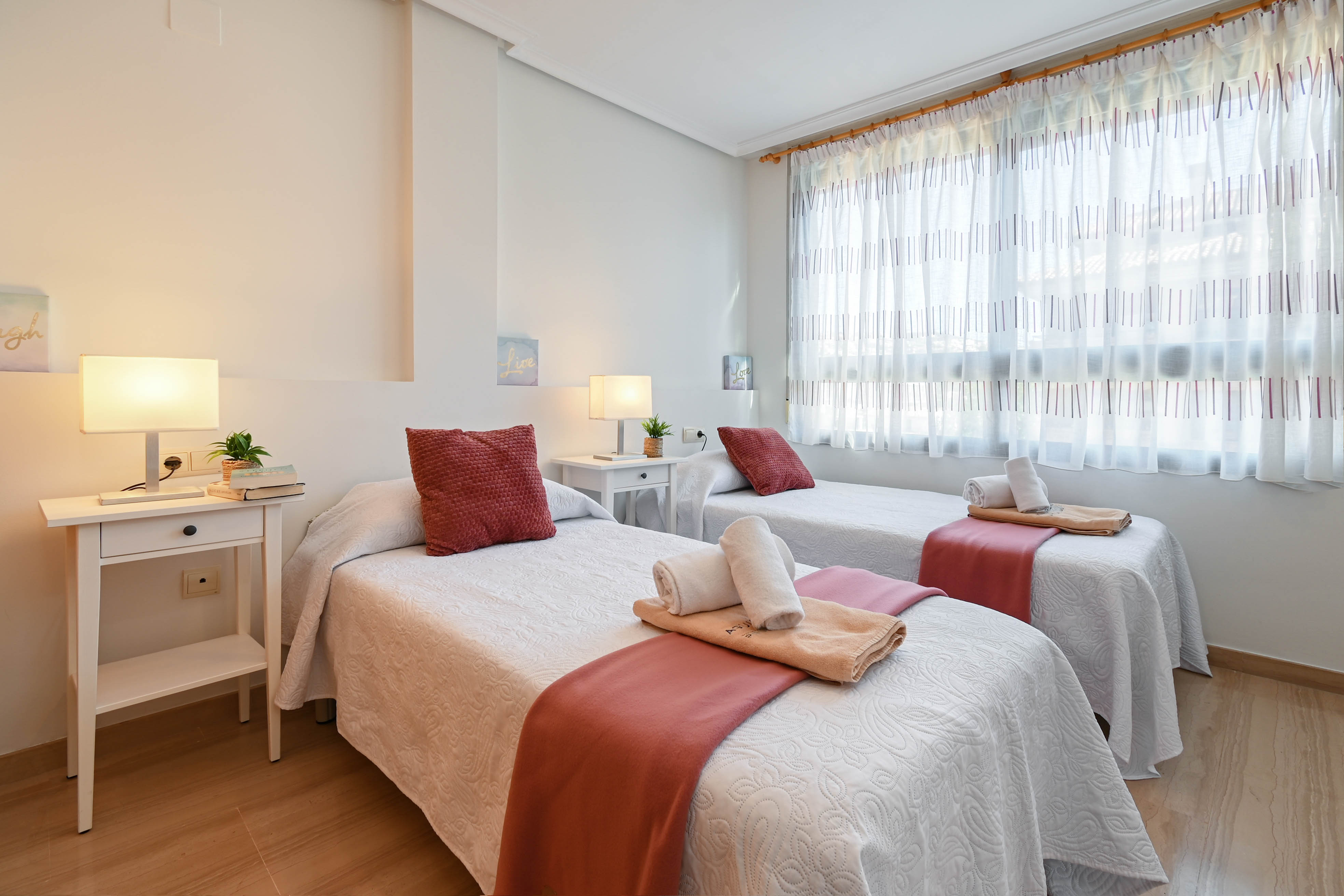 Chambre lumineuse avec deux lits simples, tables de chevet blanches et sol en bois clair. Une grande fenêtre avec rideaux translucides apporte une lumière naturelle douce.