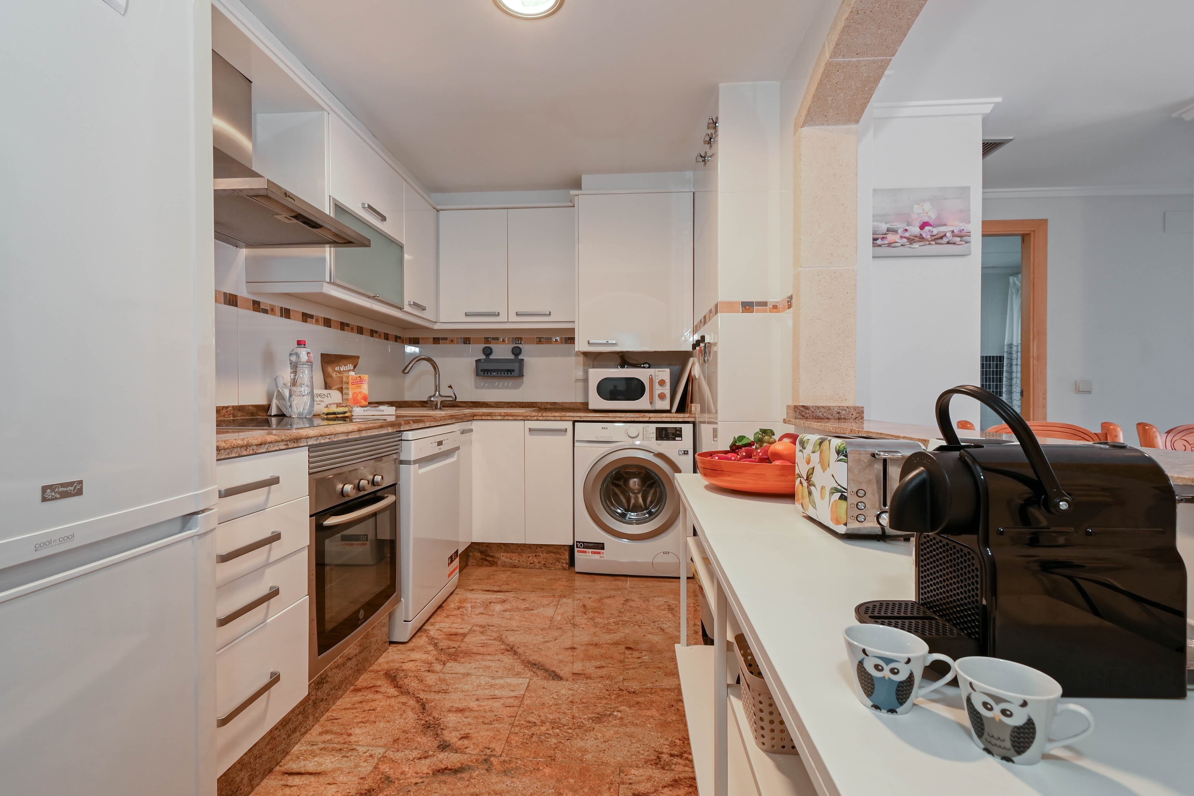 Cuisine équipée avec placards blancs, plans de travail en granit et appareils intégrés tels qu'un lave-linge, un lave-vaisselle et une machine à café.