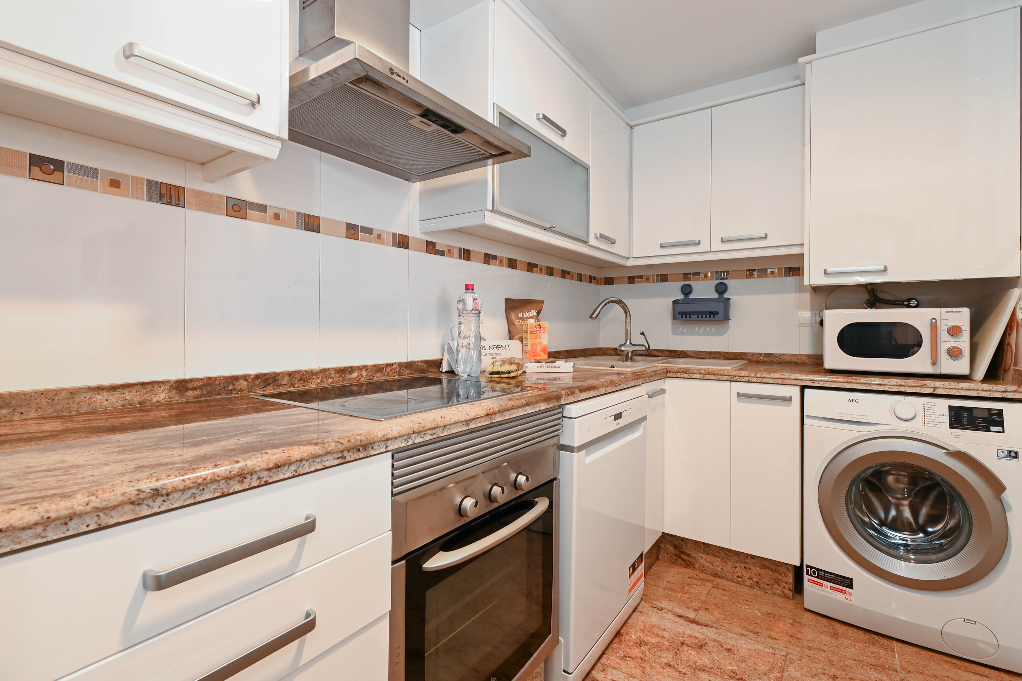 Cuisine fonctionnelle avec placards blancs, plans de travail en granit et appareils intégrés, notamment un lave-linge, un lave-vaisselle et un four en inox.