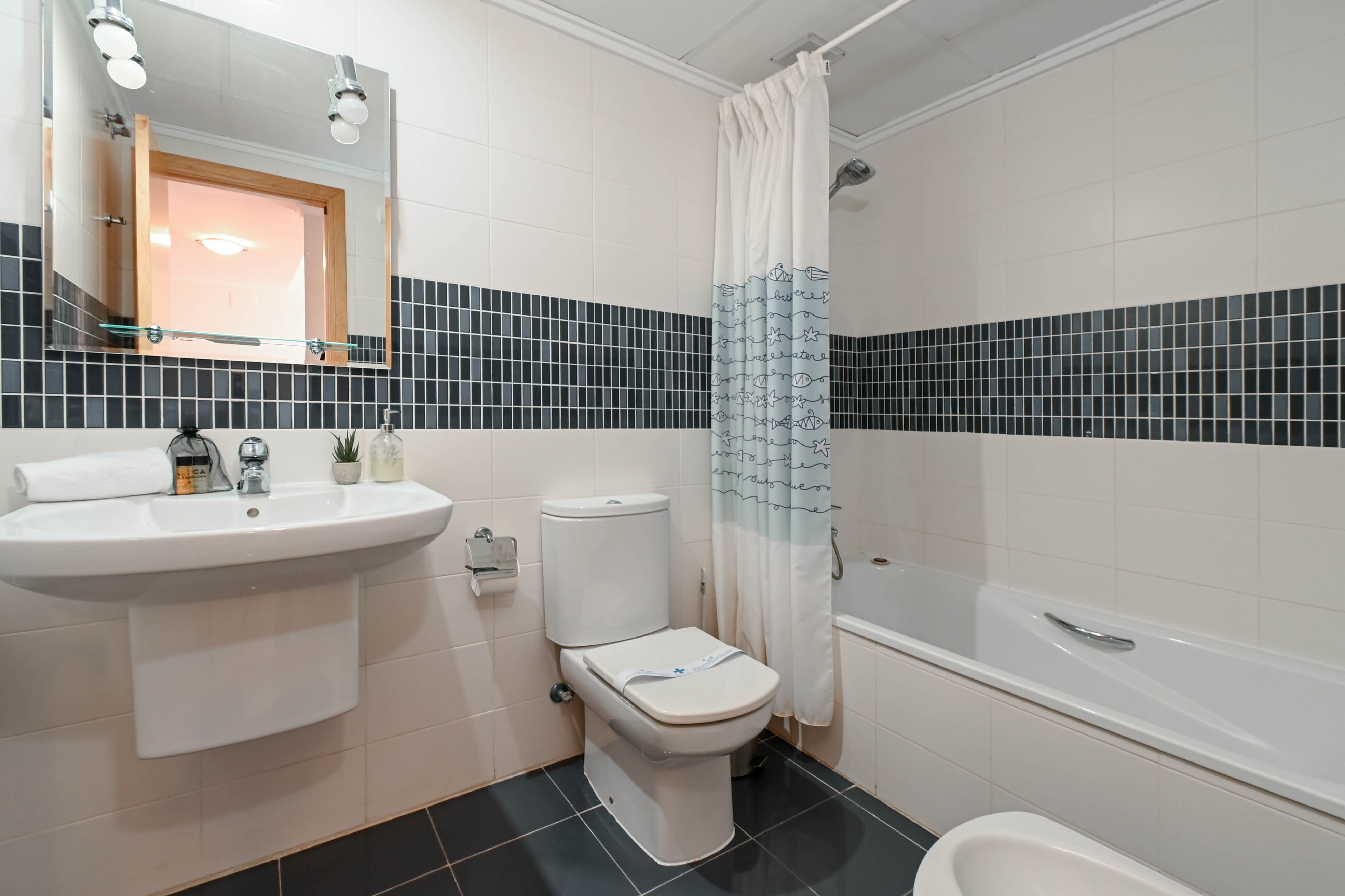 Salle de bain avec carrelage blanc et frise en mosaïque foncée, équipée d'une baignoire, d'un lavabo suspendu et d'un miroir éclairé.