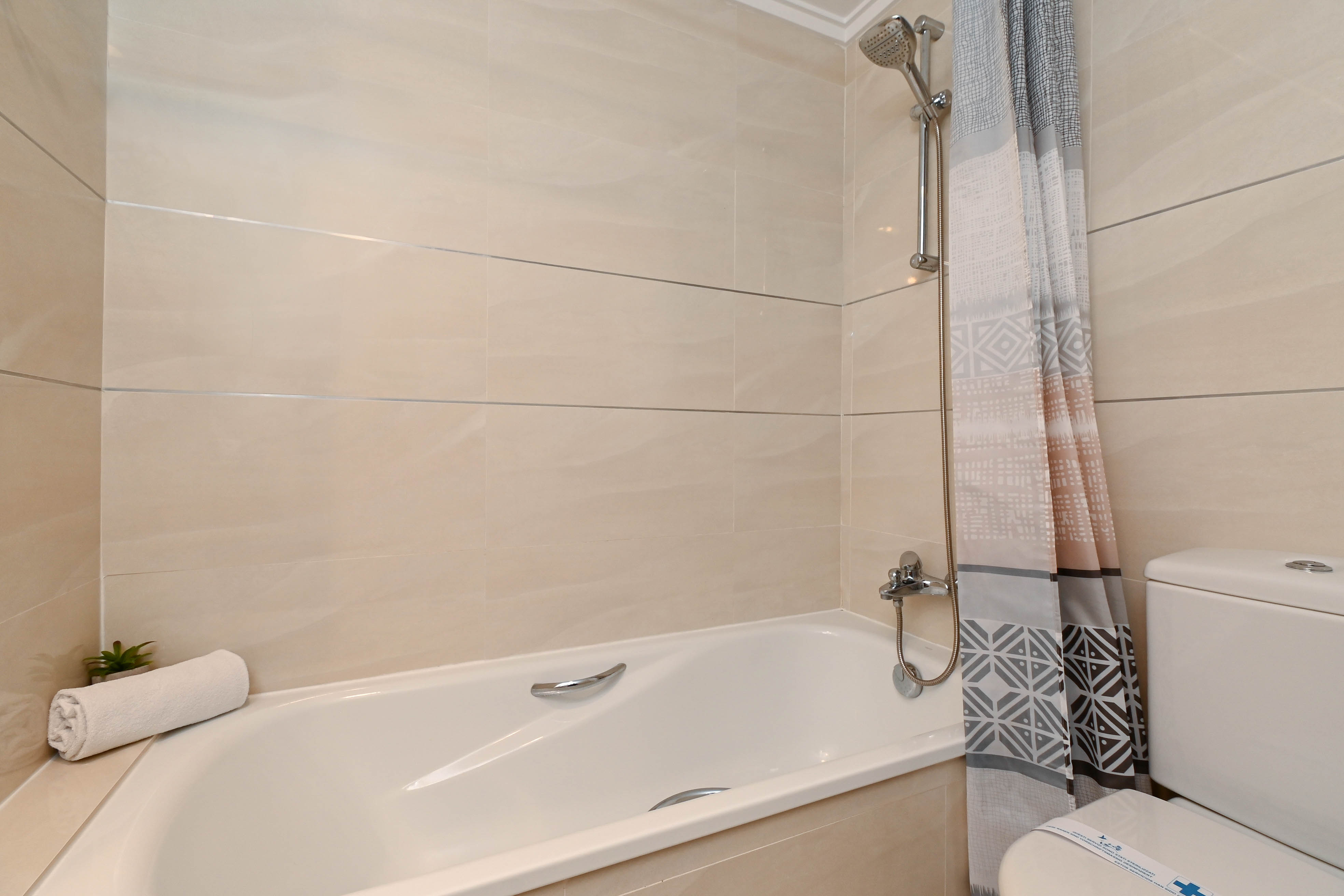 Salle de bain fonctionnelle avec baignoire, robinetterie chromée et murs en carrelage beige. Elle dispose d'un rideau de douche aux motifs géométriques.