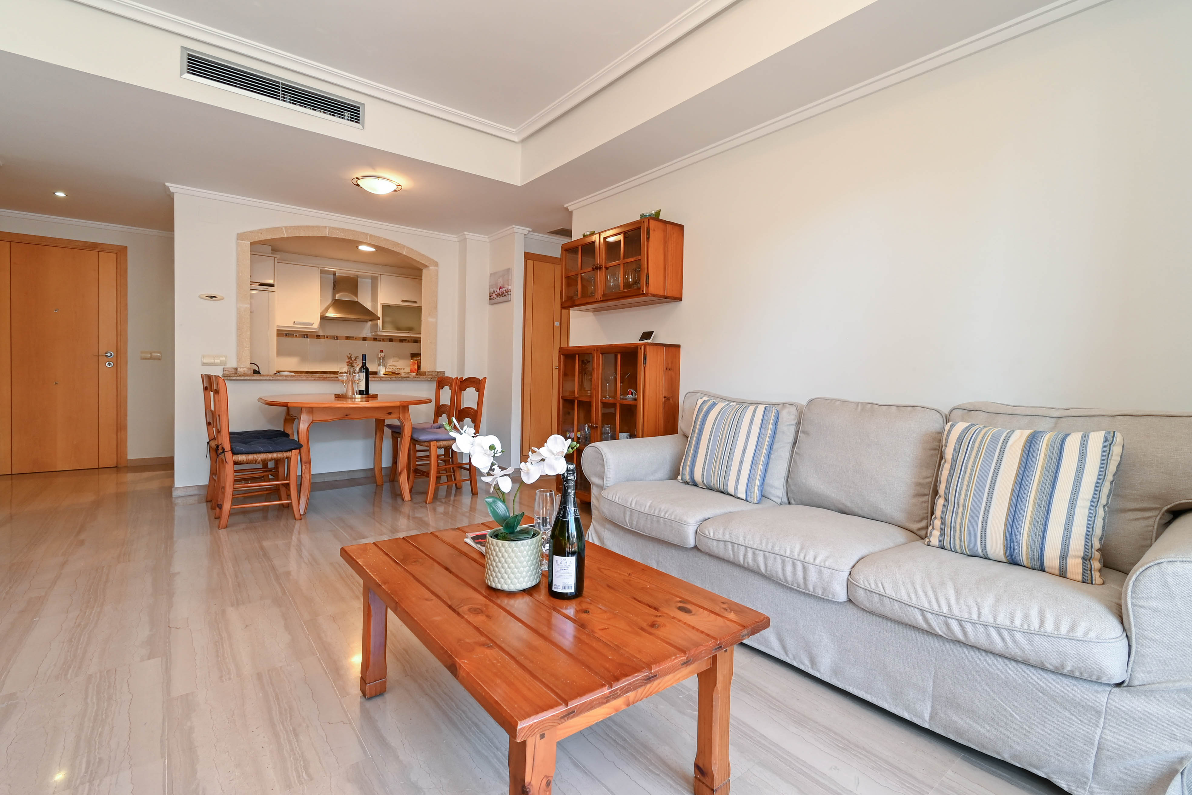 Salon ouvert avec sol en marbre, canapé beige, mobilier en bois massif et une arche en pierre ouvrant sur la cuisine équipée.