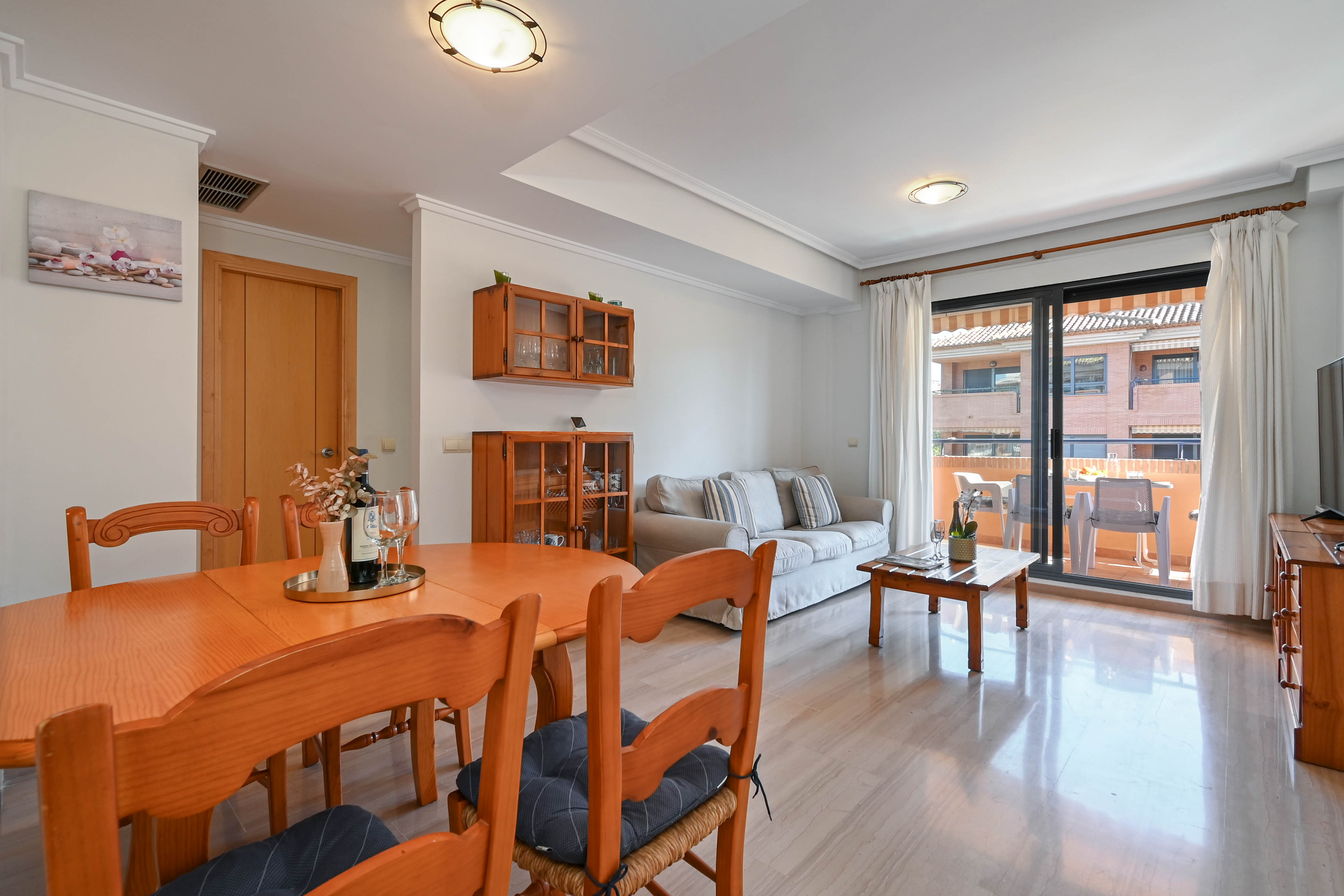 Salon-salle à manger lumineux avec mobilier en bois, sol en marbre poli et accès direct à une terrasse meublée par de grandes baies vitrées coulissantes.