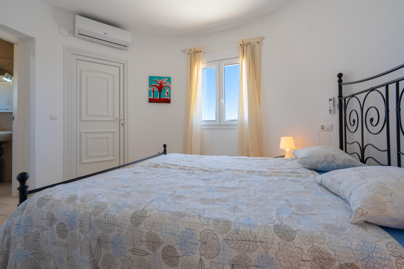 Chambre lumineuse avec cadre de lit en fer forgé noir, climatisation, fenêtre en PVC et accès direct à une salle de bain attenante.