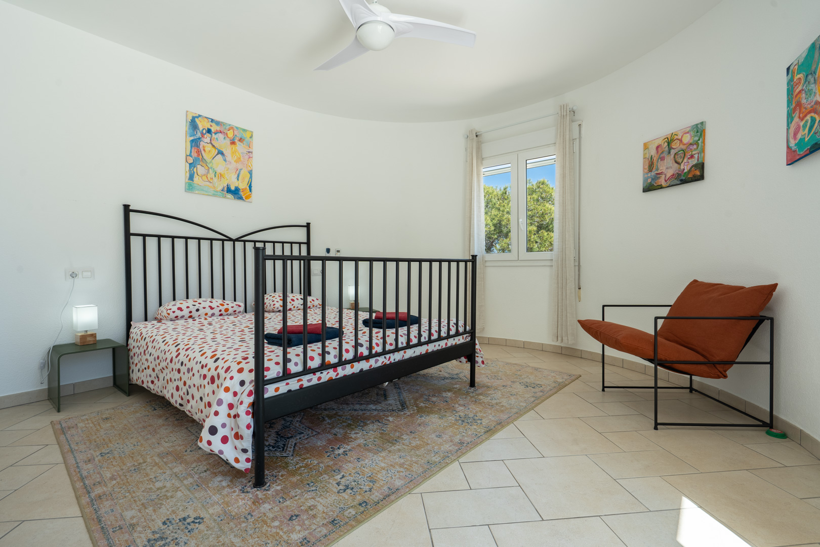 Chambre lumineuse avec mur incurvé, cadre de lit en métal noir, sol en carrelage clair et fenêtre PVC offrant une vue sur la végétation.