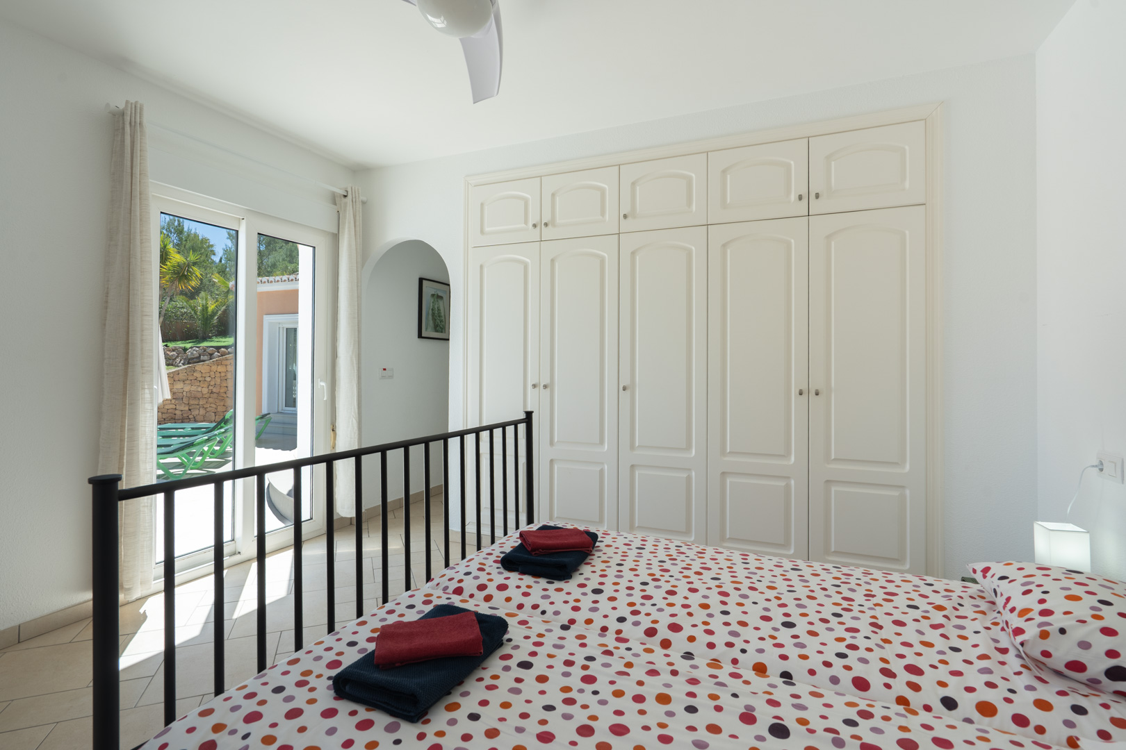Chambre lumineuse avec placards intégrés blancs, ventilateur de plafond et accès direct à la terrasse par des baies vitrées en PVC.