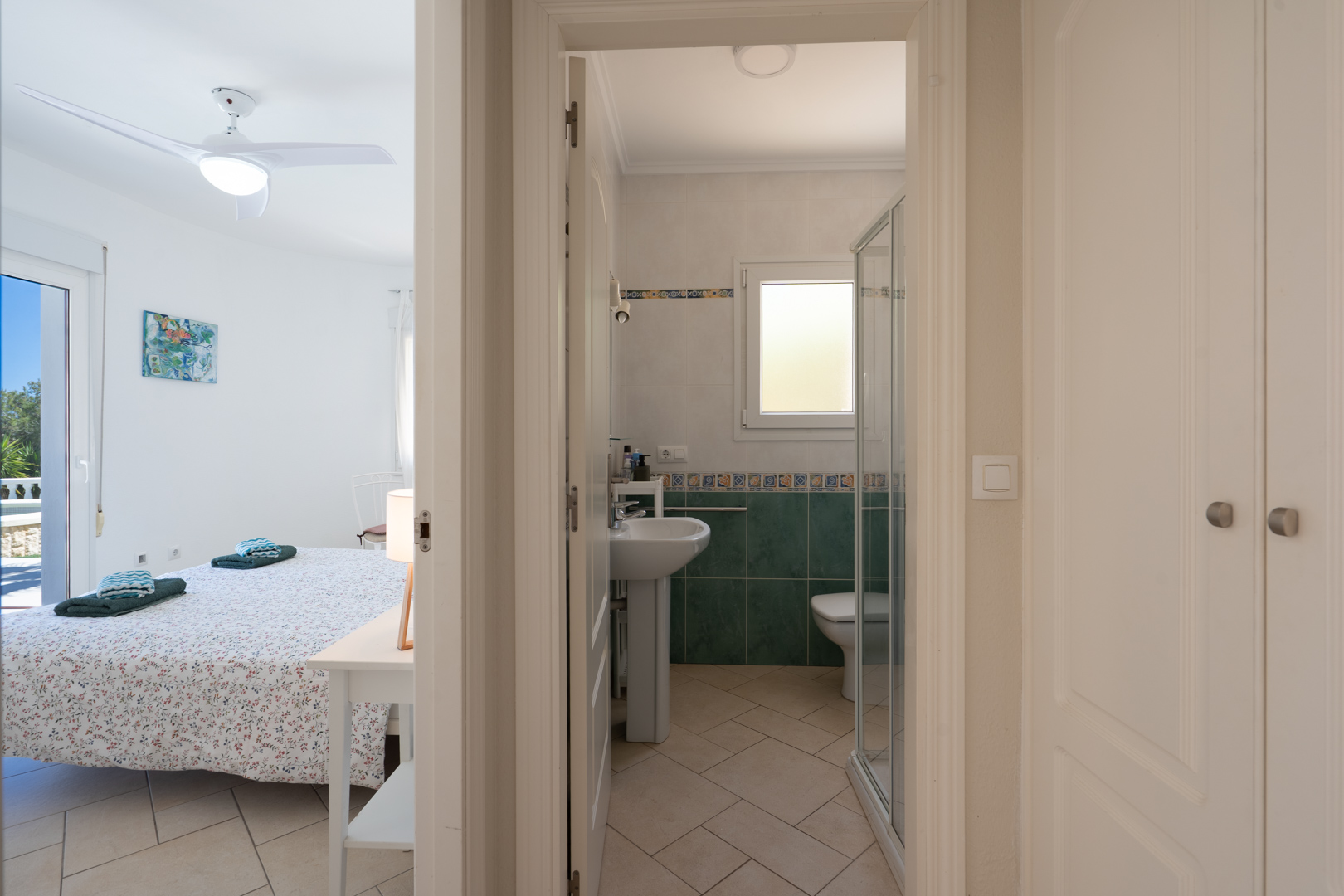 Chambre lumineuse avec ventilateur de plafond et accès extérieur, située à côté d'une salle de bain avec douche et de placards intégrés dans le couloir.