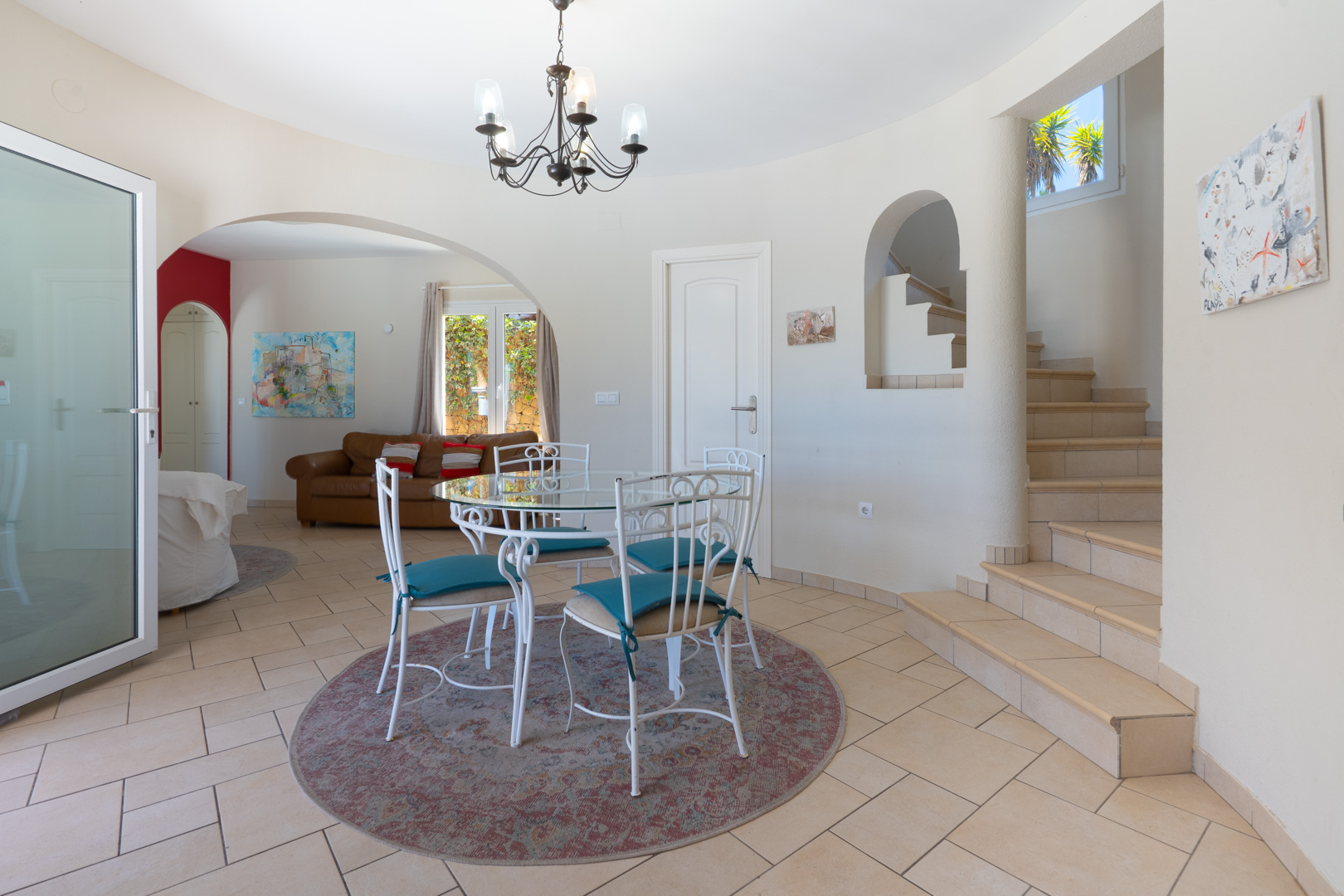 Coin repas avec table en verre et chaises en fer forgé blanc. L'espace s'ouvre sur un escalier carrelé et un salon adjacent via une arche élégante.