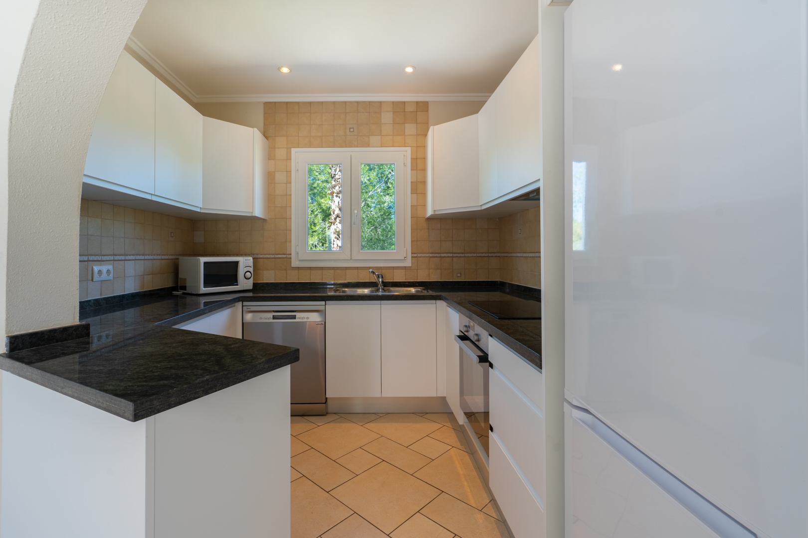 Cuisine fonctionnelle en U avec plans de travail en granit sombre, meubles blancs, électroménagers intégrés et une fenêtre en PVC offrant une vue sur l'extérieur.
