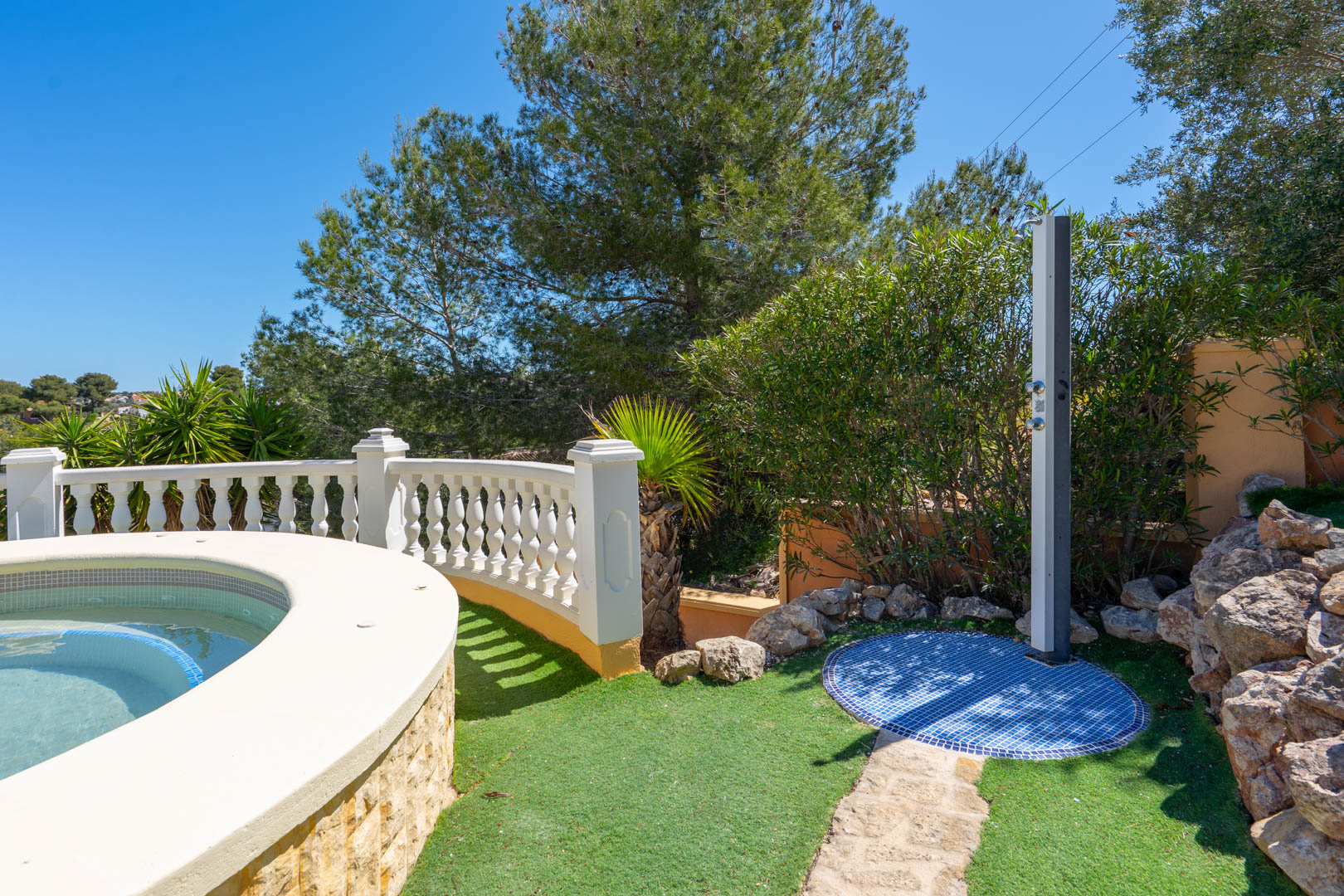 Douche extérieure avec base en mosaïque bleue, située près de la piscine à bord arrondi et entourée d'une balustrade blanche et de verdure.