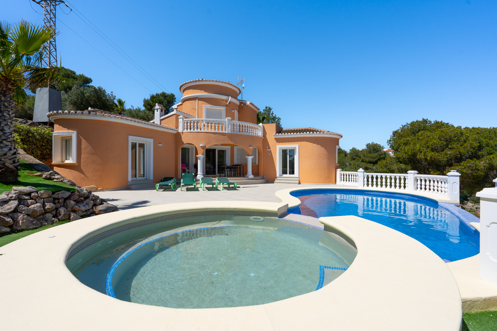 Extérieur de la villa avec piscine aux formes arrondies, vaste terrasse ensoleillée et façade méditerranéenne aux tons terre cuite et balustrades blanches.