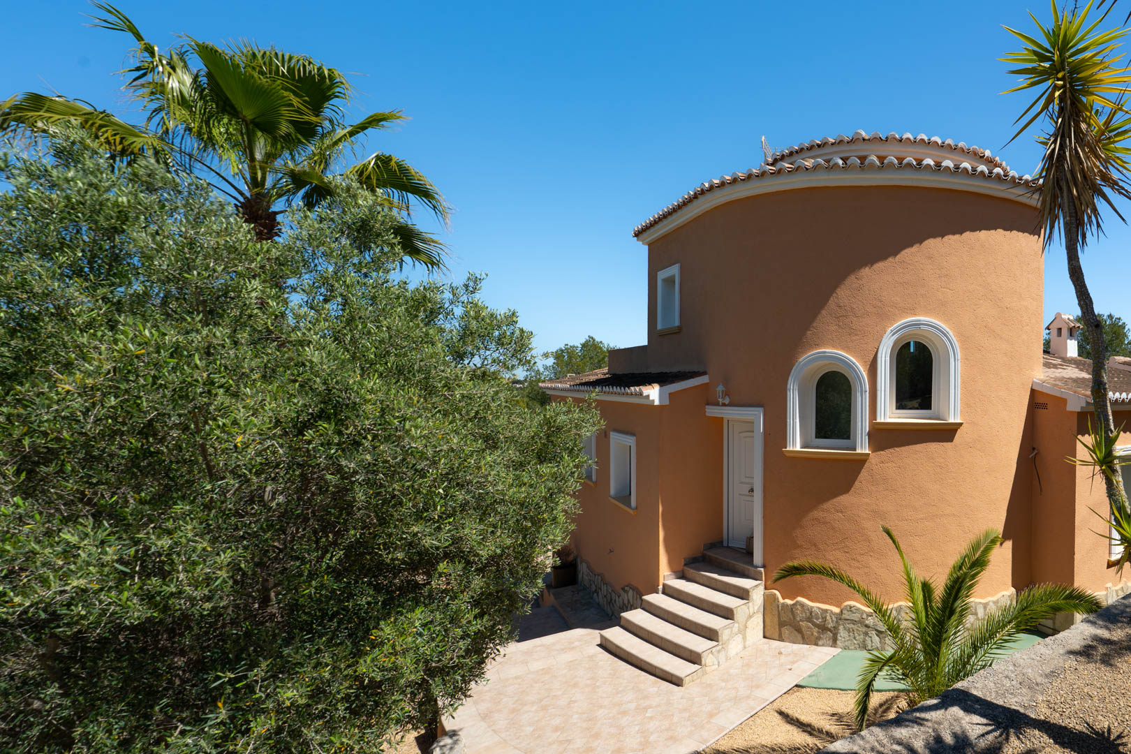Extérieur de la villa avec une façade ocre, une tour circulaire aux fenêtres cintrées et un escalier en pierre menant à l'entrée principale.