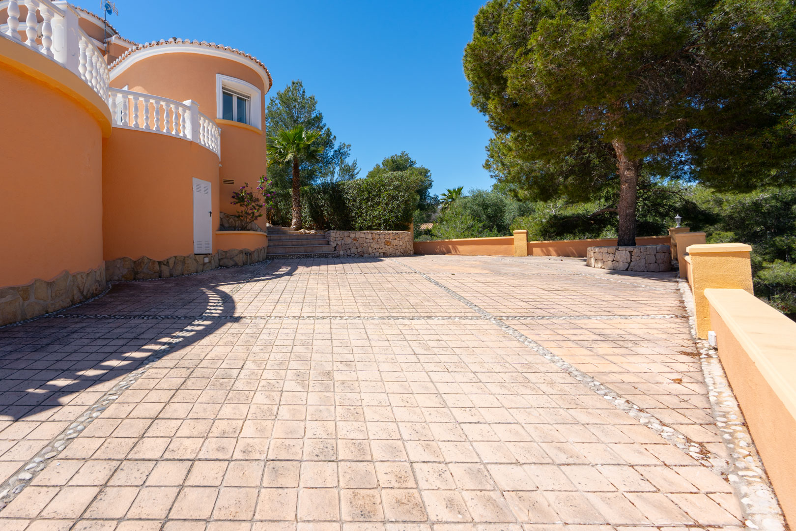 Grande terrasse pavée avec bordures en galets, murs courbes de couleur terre cuite et balustrades blanches, entourée de pins et de végétation locale.