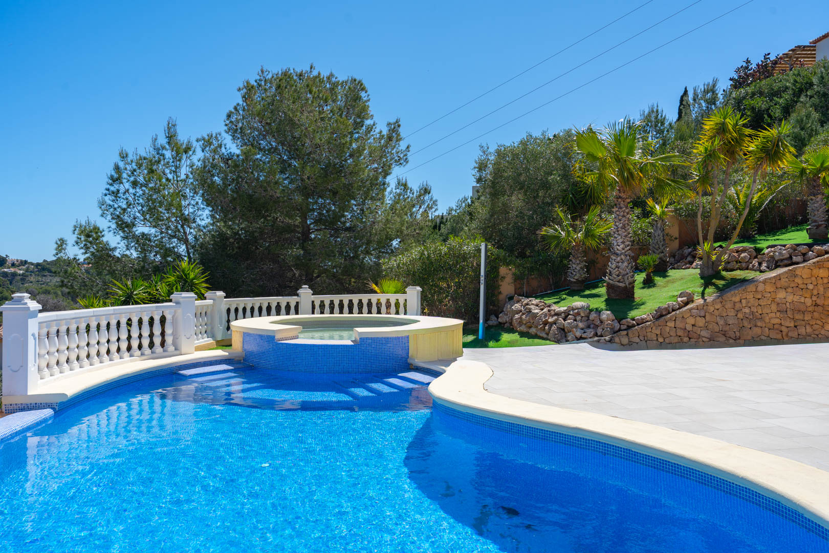 Piscine de 10x5 m avec spa circulaire intégré, carrelage en mosaïque bleue, balustrade en pierre blanche et jardin paysager avec palmiers.