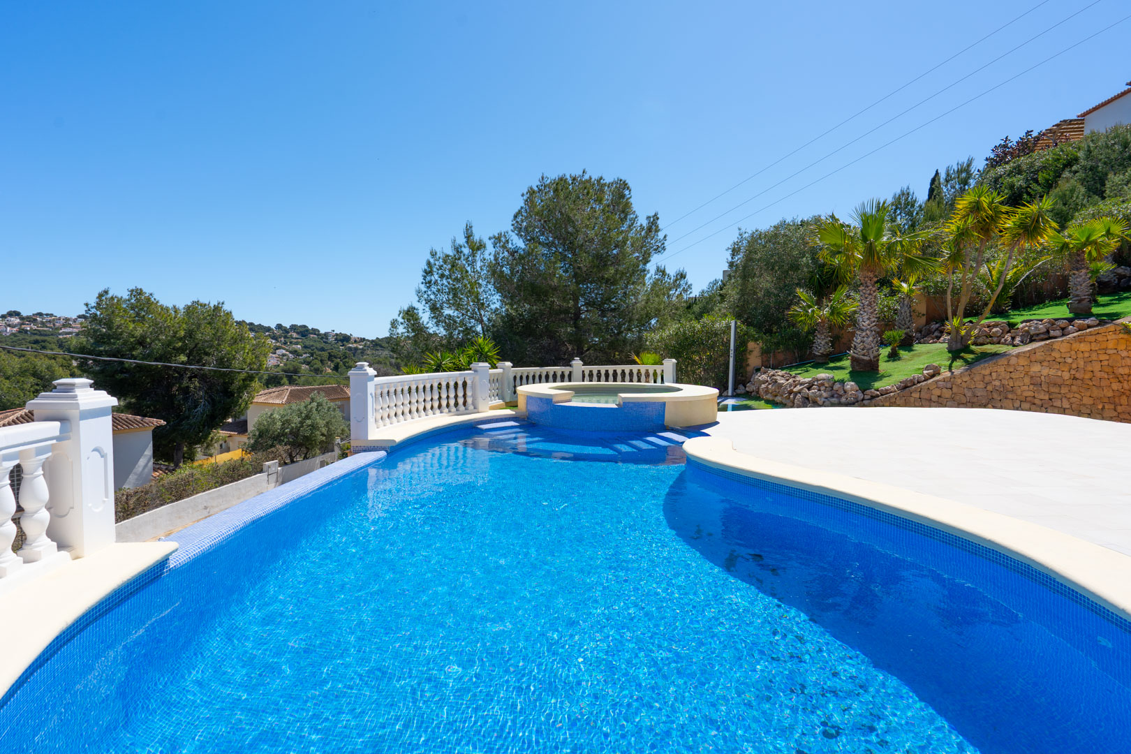Piscine en mosaïque bleue avec jacuzzi intégré, entourée d'une terrasse pavée, d'une balustrade blanche et d'un jardin paysager avec palmiers.