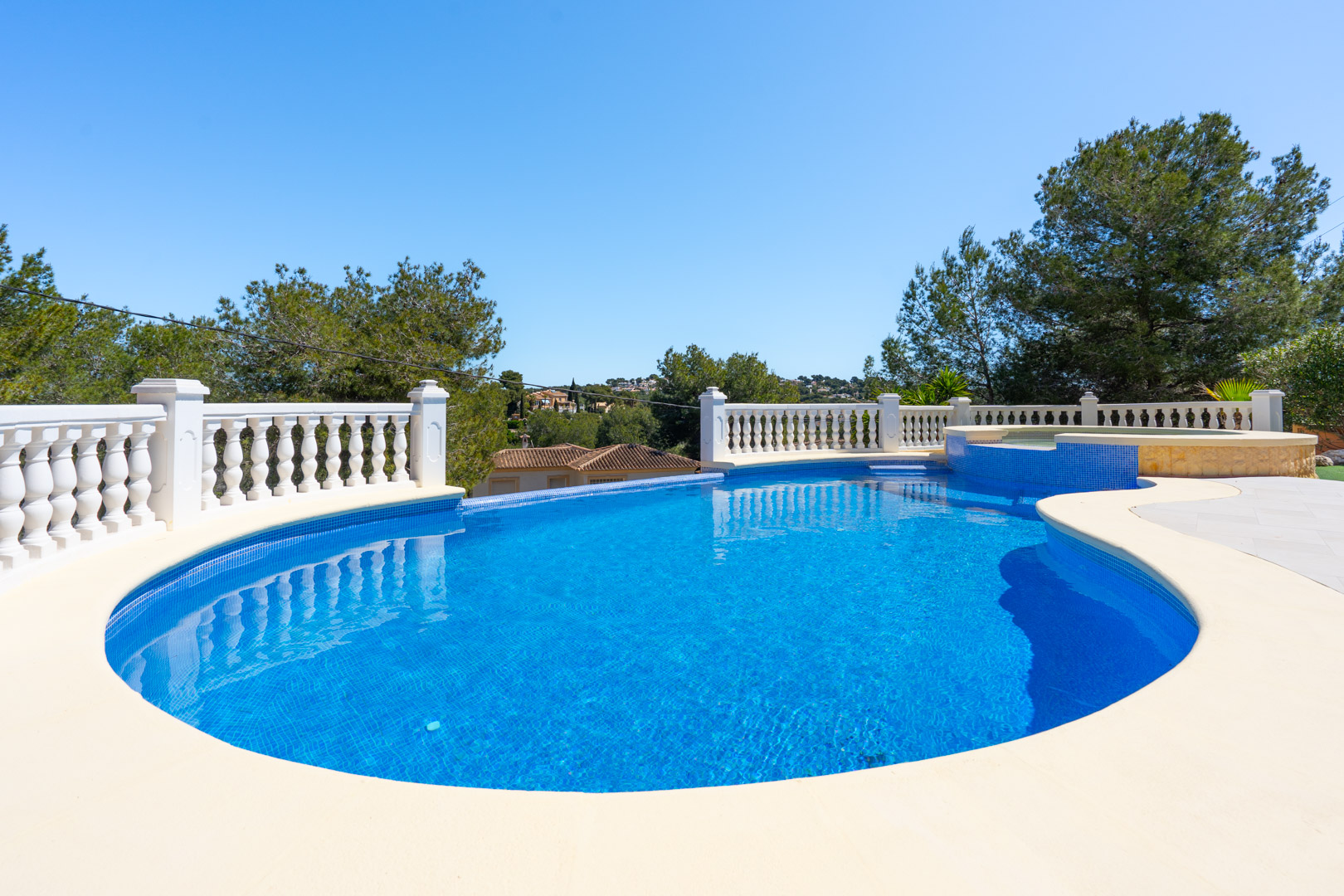 Piscine extérieure de forme libre avec mosaïque bleue, entourée d'une balustrade blanche classique et d'une terrasse offrant une vue dégagée sur la pinède.