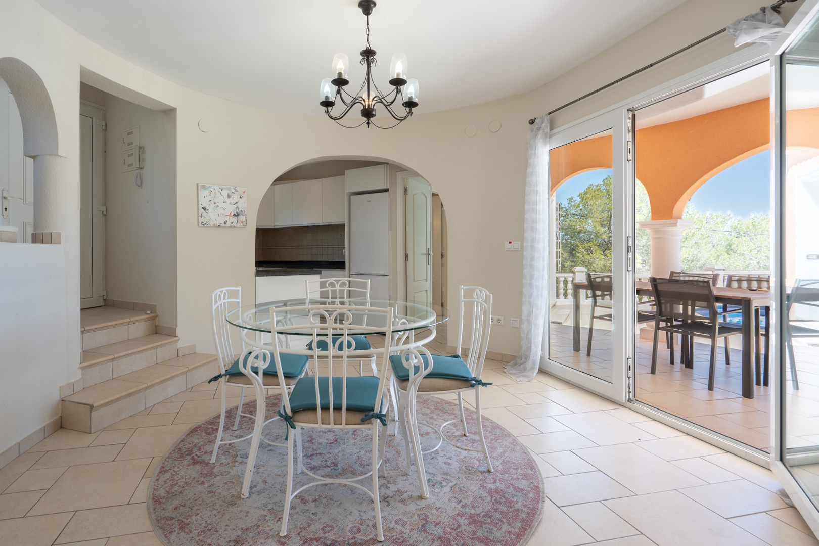 Salle à manger lumineuse avec table en verre et chaises en fer forgé, offrant un accès à la cuisine par une arche et une ouverture sur la terrasse.
