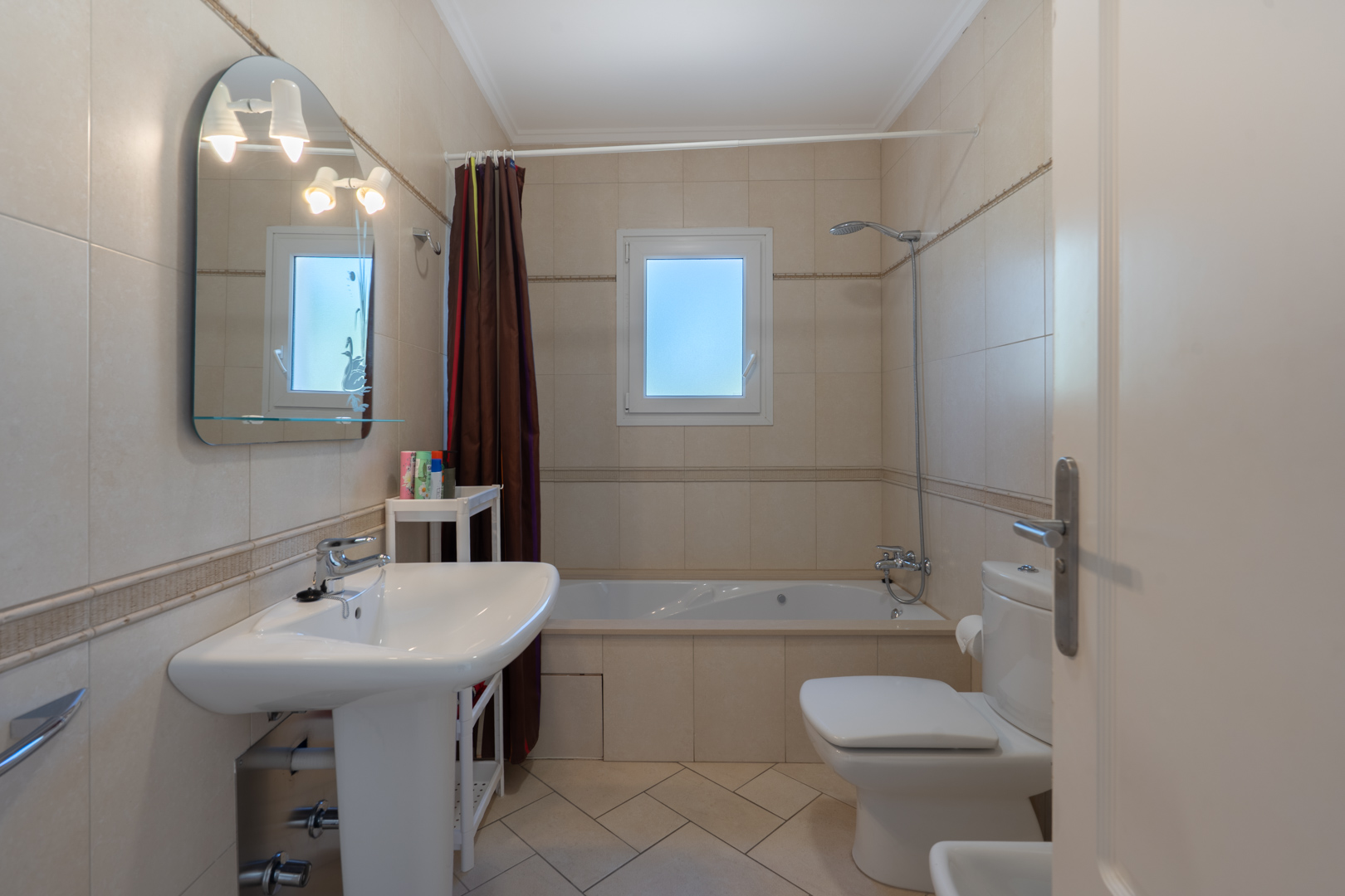 Salle de bain avec carrelage beige, baignoire encastrée, lavabo sur colonne et miroir cintré avec éclairage intégré près d'une fenêtre en PVC.