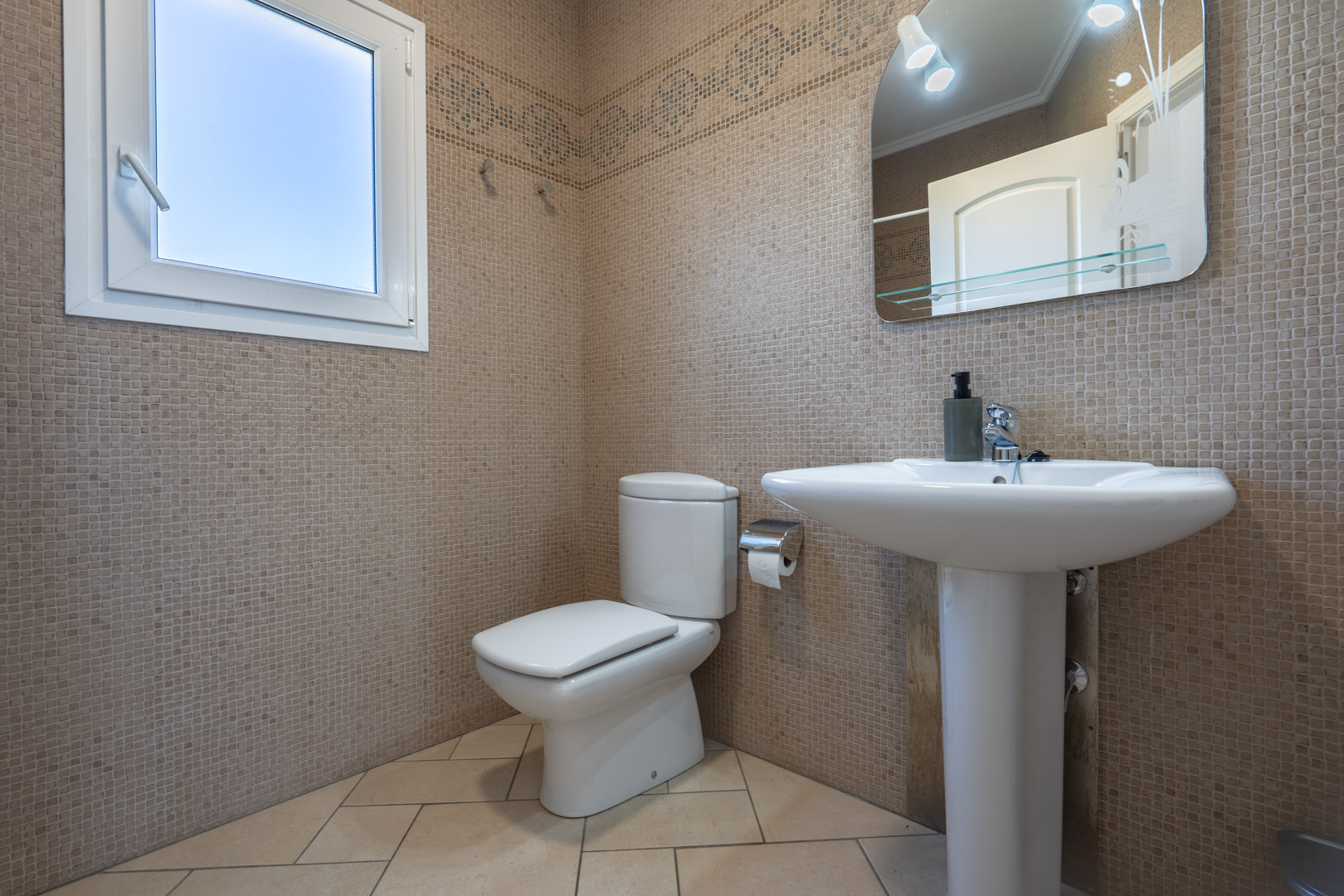 Salle de bain avec murs en mosaïque beige, lavabo sur colonne et fenêtre en PVC blanc offrant une luminosité naturelle.