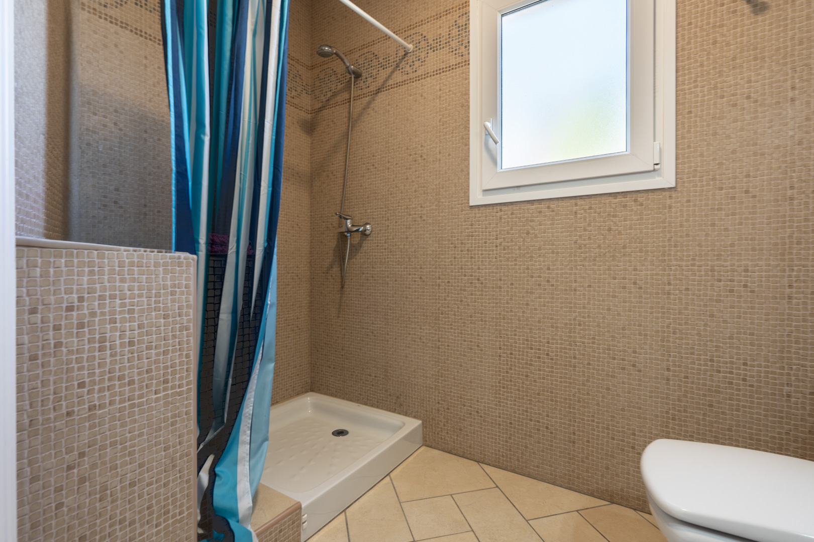 Salle de bain avec murs en mosaïque brune, receveur de douche blanc avec rideau rayé et fenêtre en PVC blanc offrant une lumière naturelle diffuse.