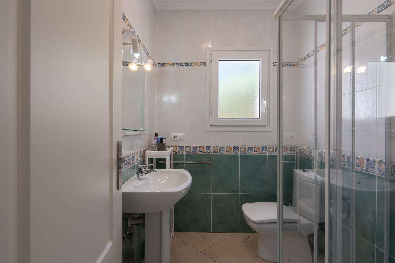 Salle de bain fonctionnelle avec carrelage vert et blanc, lavabo sur colonne, douche vitrée et fenêtre en PVC à verre dépoli.