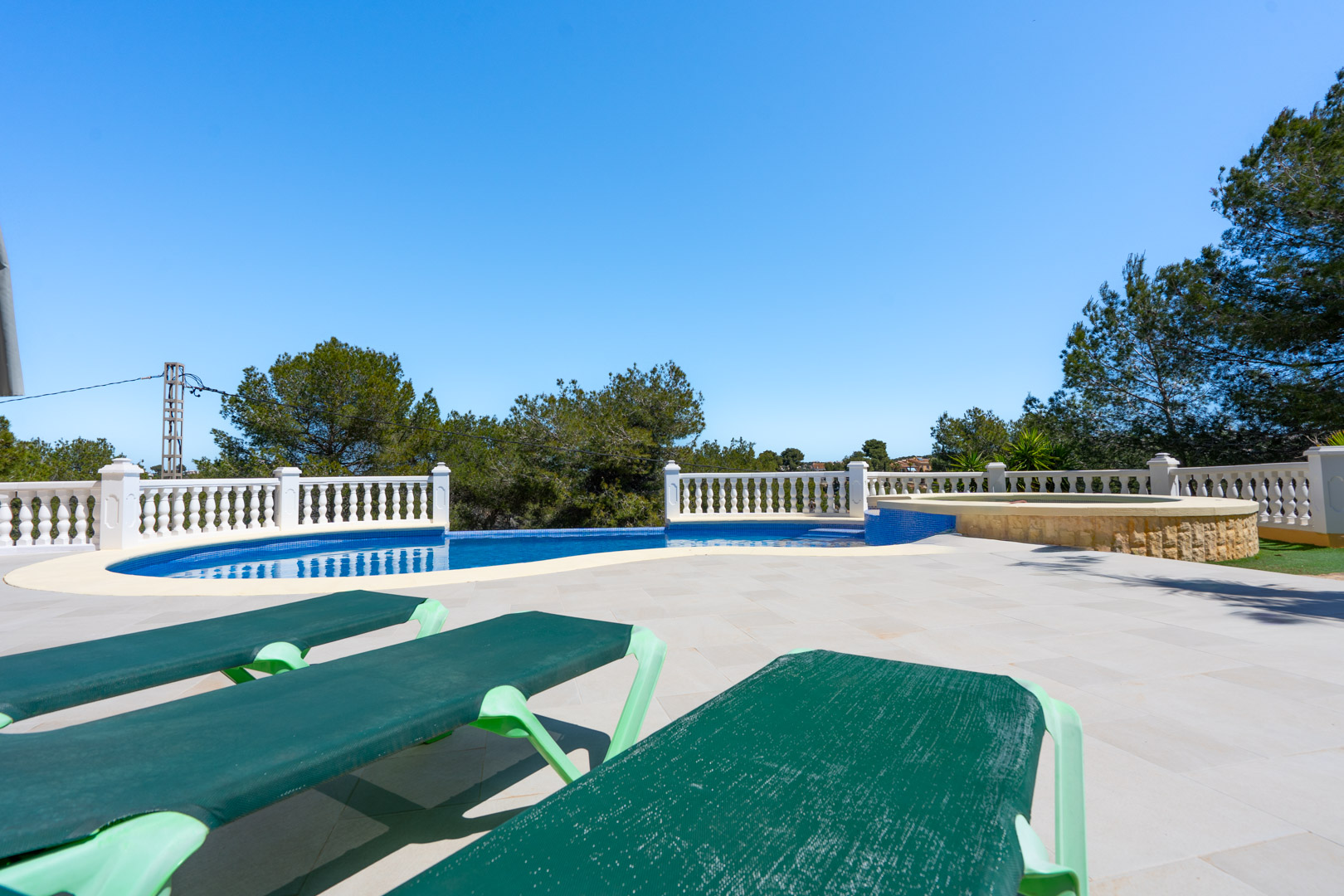 Terrasse extérieure avec piscine carrelée en bleu, balustrade blanche et jacuzzi intégré en pierre, entourée de pins méditerranéens et de chaises longues.