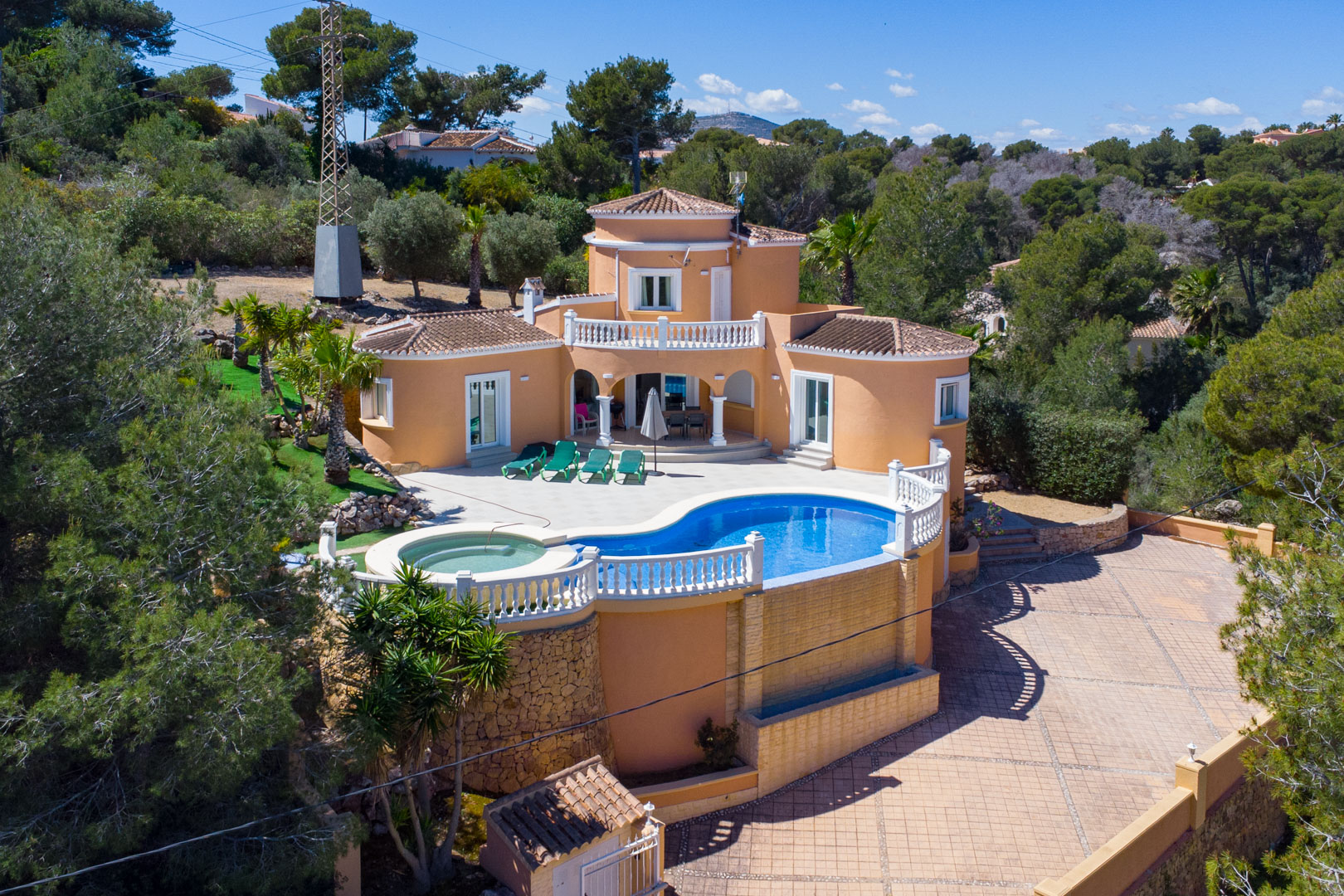 Vue aérienne de la villa méditerranéenne avec piscine courbe, jacuzzi intégré et balustrades blanches, entourée de pins et d'une grande cour pavée.