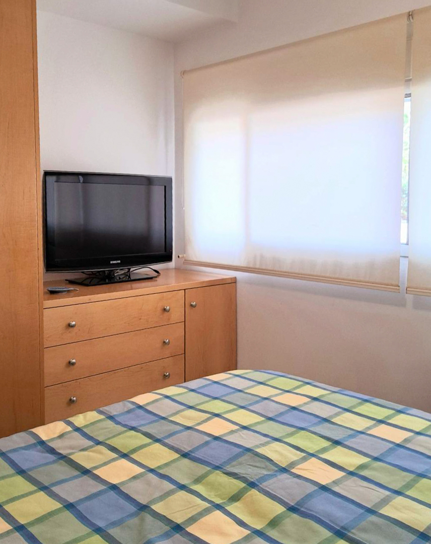 Chambre avec mobilier en bois sur mesure, télévision sur commode et fenêtre équipée de stores enrouleurs laissant passer la lumière.