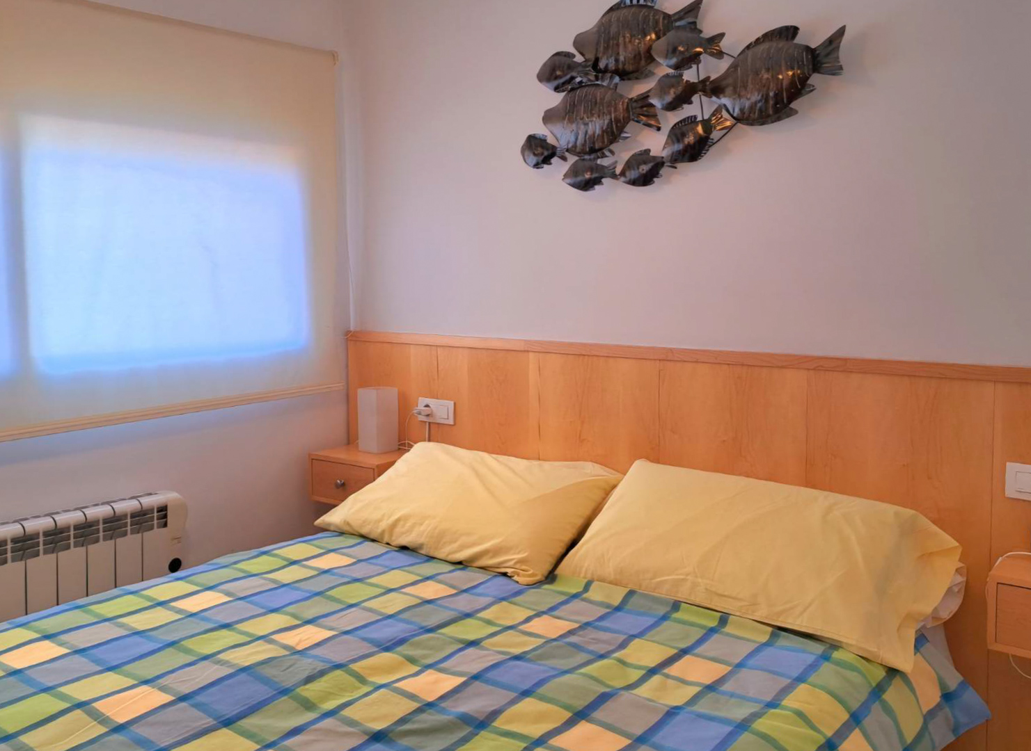 Chambre avec tête de lit intégrée en bois, tables de chevet assorties et décoration murale en métal. Comprend un radiateur électrique et une fenêtre avec store.