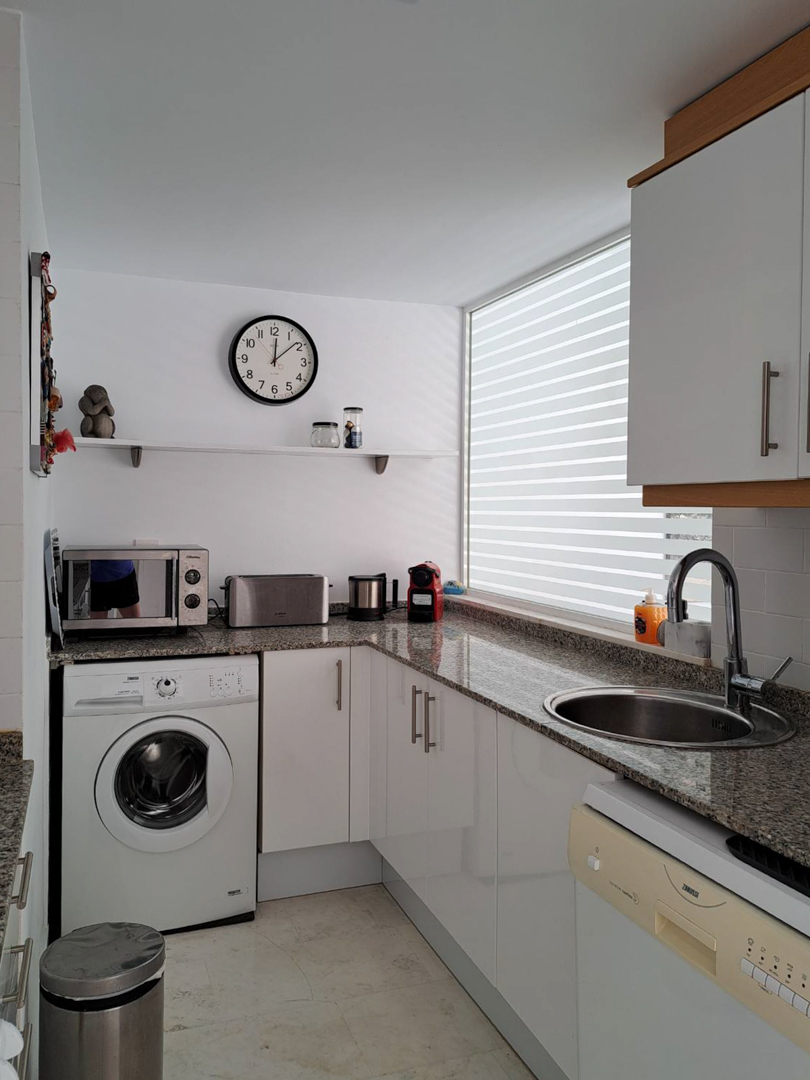 Cuisine fonctionnelle en L avec plan de travail en granit, meubles blancs, lave-linge intégré et une grande fenêtre apportant une luminosité naturelle.